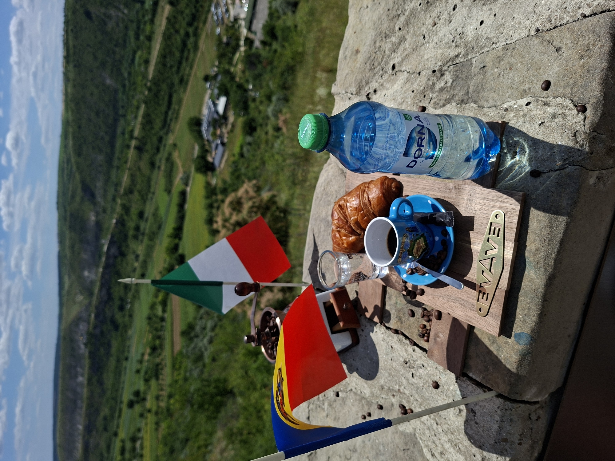 A close-up shot captures a breakfast setting on a stone ledge overlooking a lush valley in Trebujeni, Moldova. Two small flags, one Italian and one Moldovan, are prominently displayed. In the center of the composition is a small wooden tray holding a cup of black coffee, a croissant, a glass of water, and scattered coffee beans. To the right, a blue plastic bottle of "DORNA" mineral water stands upright. The background reveals a vibrant green landscape with rolling hills and a clear blue sky dotted with white clouds. The focus is sharp on the breakfast items and flags, with the background softly blurred.