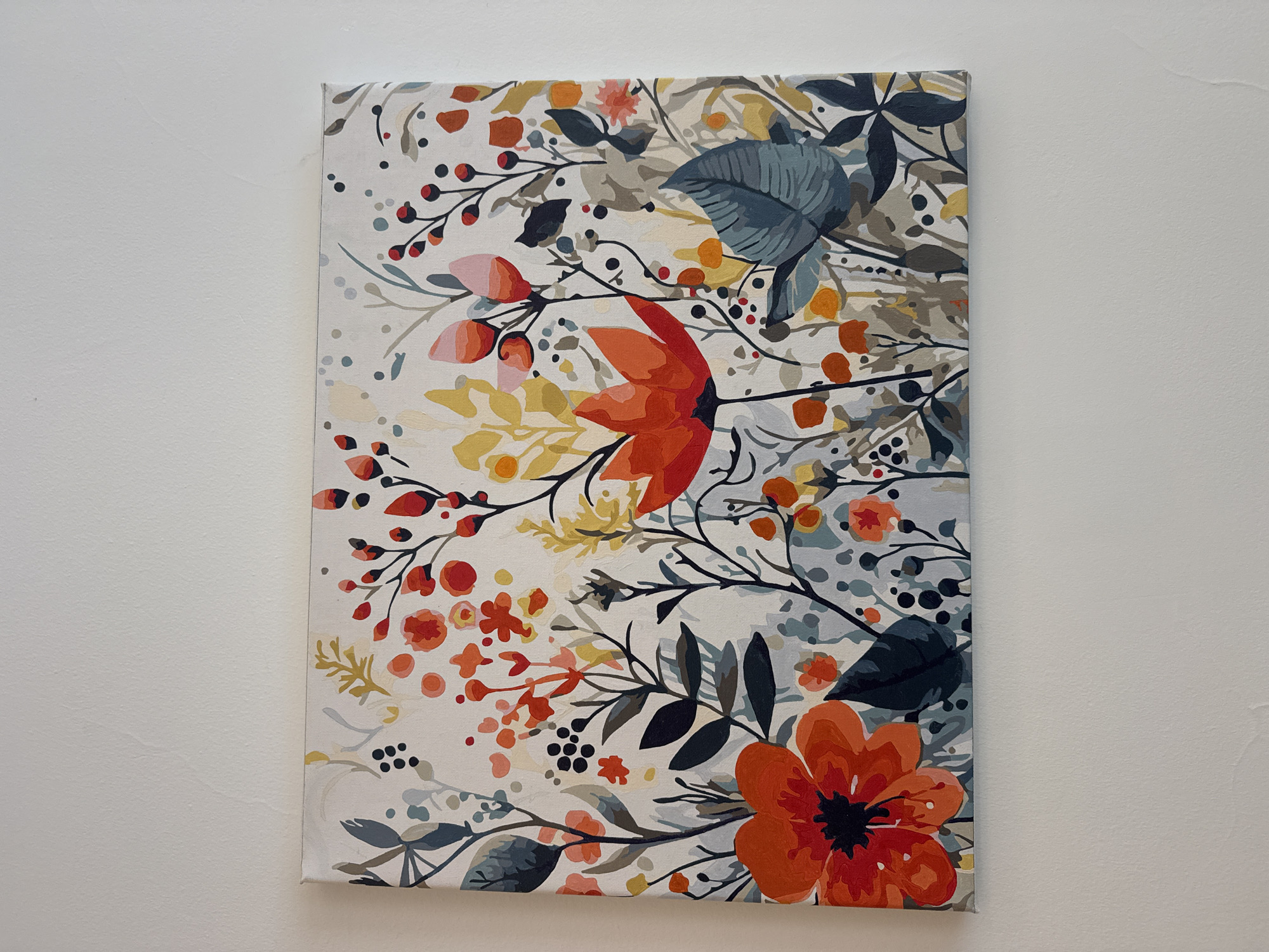 The image shows a rectangular painting depicting a floral design, hung on a white wall. The painting features stylized flowers, leaves, and berries in shades of orange, red, yellow, gray, and blue. The background of the painting is a light beige. The painting is mounted on a canvas frame. The wall is plain and white with slight texture. There is no visible information to indicate that the photo was taken in Pattaya City, Thailand.
