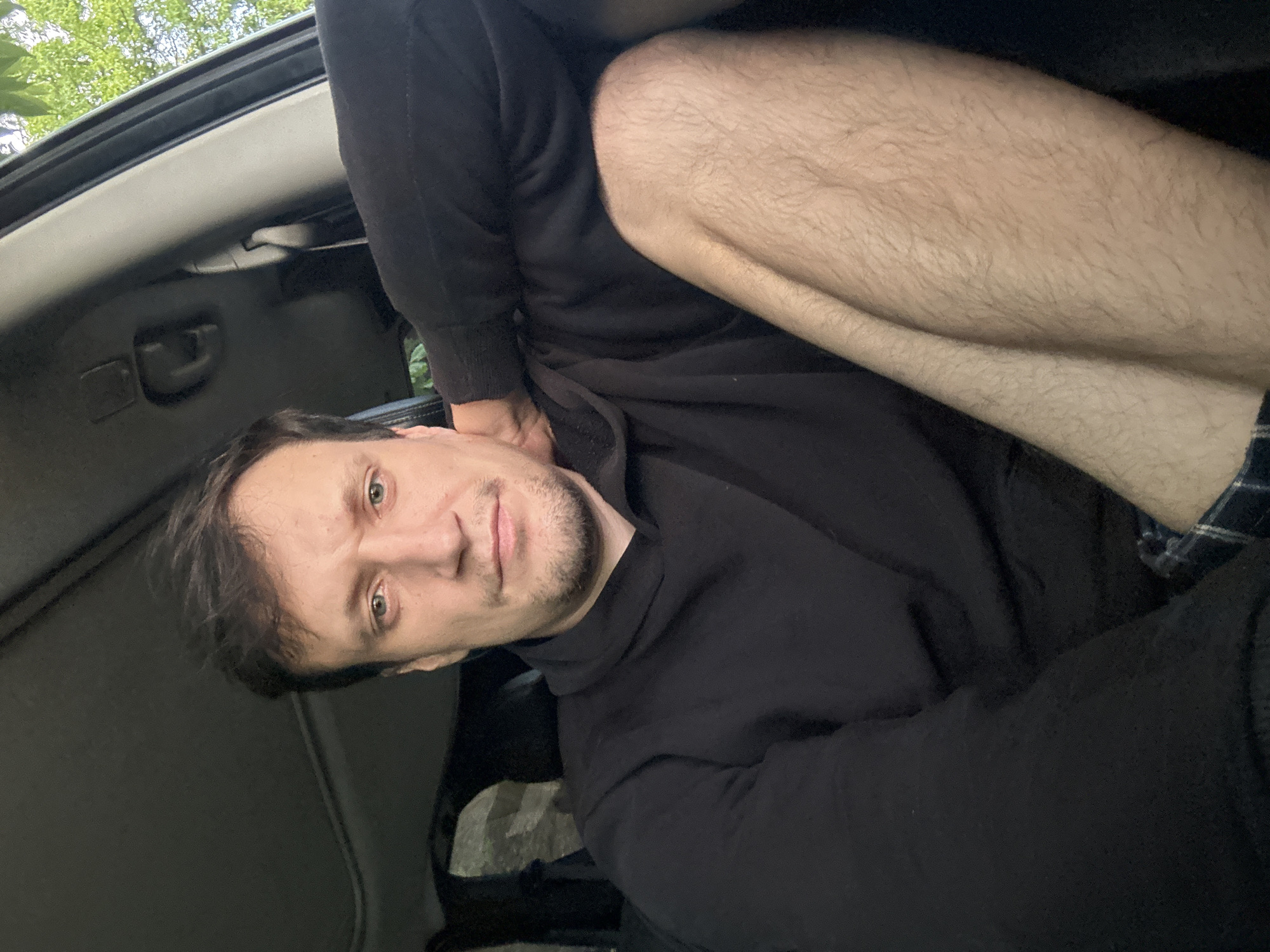 A man with medium brown hair and a beard sits in the driver's seat of a vehicle. He is wearing a black hooded sweatshirt and plaid shorts. His left arm rests on the armrest, and his right hand is behind his head. The vehicle's sunroof is visible in the background, as is a tree outside the open window. The image appears to have been taken during the day. The location is Makiv, Ukraine.