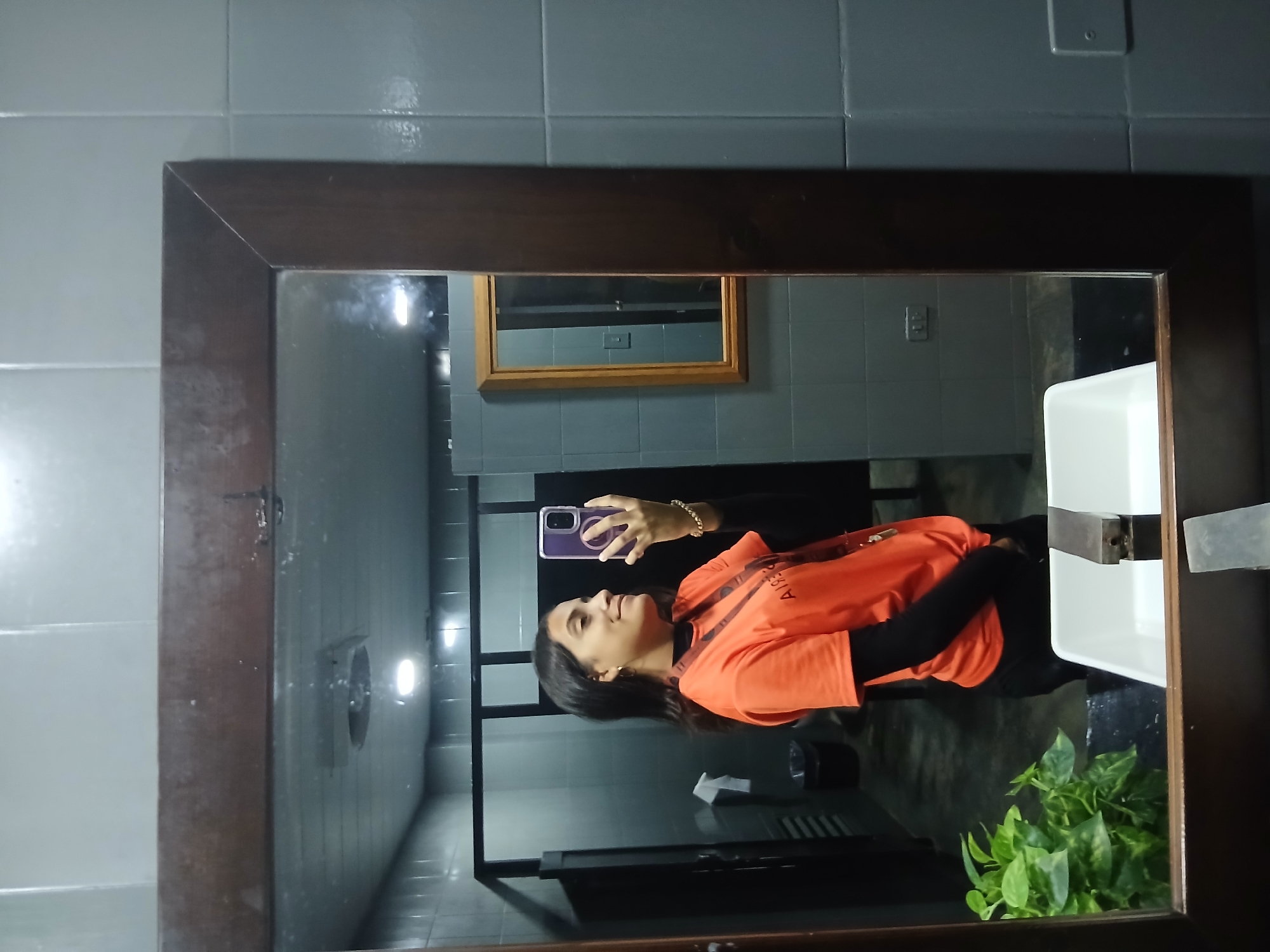A young woman with long dark hair is taking a mirror selfie in a public restroom. She is wearing an orange t-shirt over a black long-sleeved shirt and black pants. She is holding a purple smartphone in her right hand and has a bracelet on her left wrist. She is smiling and looking at her reflection. The restroom has gray tiled walls and a wooden framed mirror. In the reflection, other stalls and a sink with a plant are visible. The image is taken in the city of Playa El Agua, Venezuela.