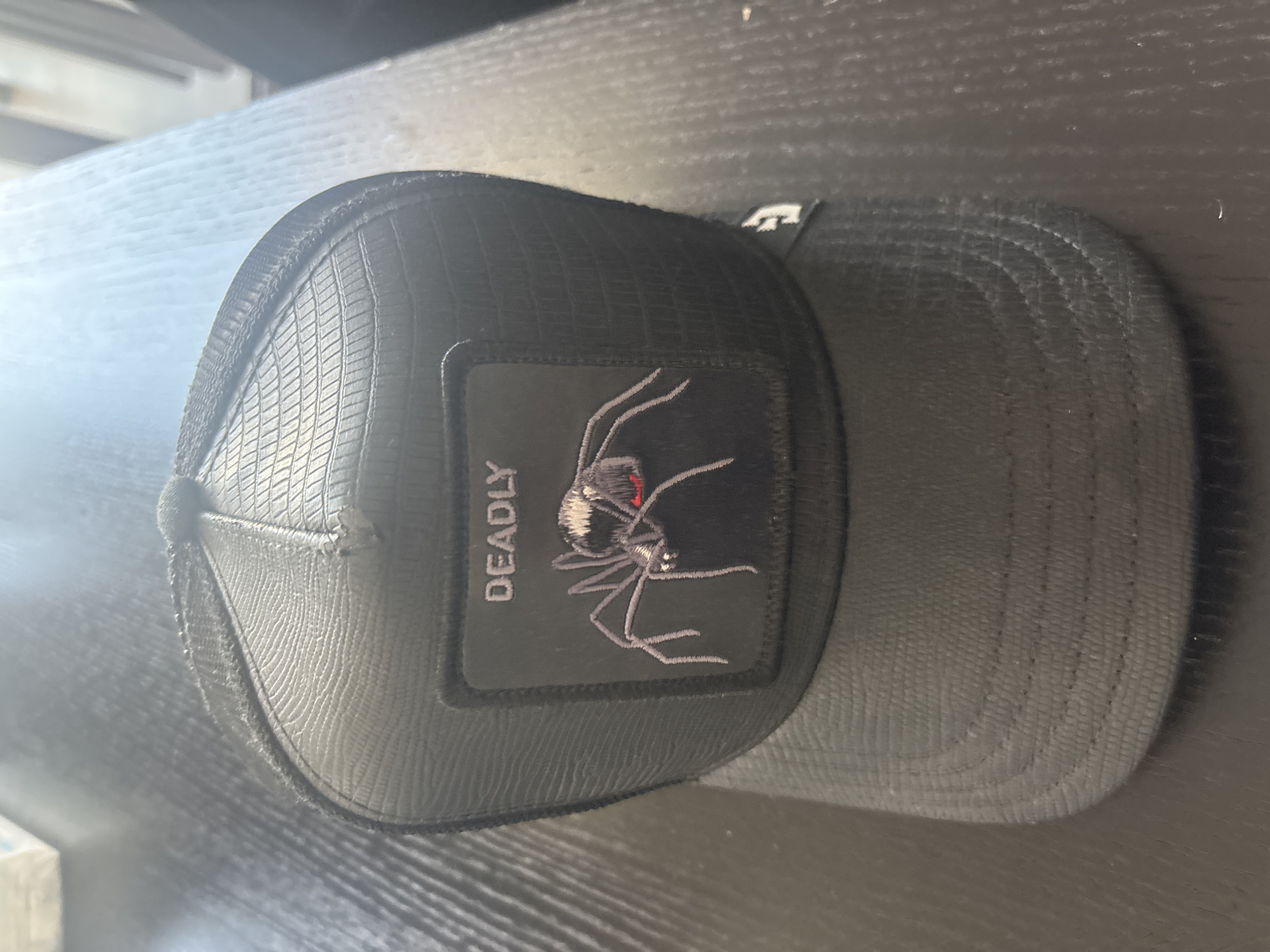 A black trucker hat with a patch featuring a spider and the word "DEADLY" is displayed on a dark, textured surface, possibly a table. The hat has a mesh back and a textured front panel. A small white logo is visible on the side of the hat. The lighting suggests an indoor setting. The image does not contain any information to suggest it is located in Dubai, United Arab Emirates.