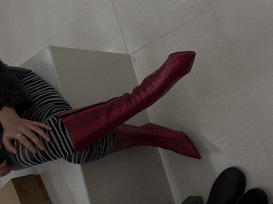 A high-angle shot features a person wearing red thigh-high, pointed-toe boots. The person is also wearing zebra-striped tights. The boots are partially resting on a light-colored ledge. A hand with a ring is visible, resting on the leg. In the bottom left corner, part of a brown box with a Nike logo can be seen. Near the bottom right, two black shoes are partially visible. The background is a light-colored, possibly tiled, surface.