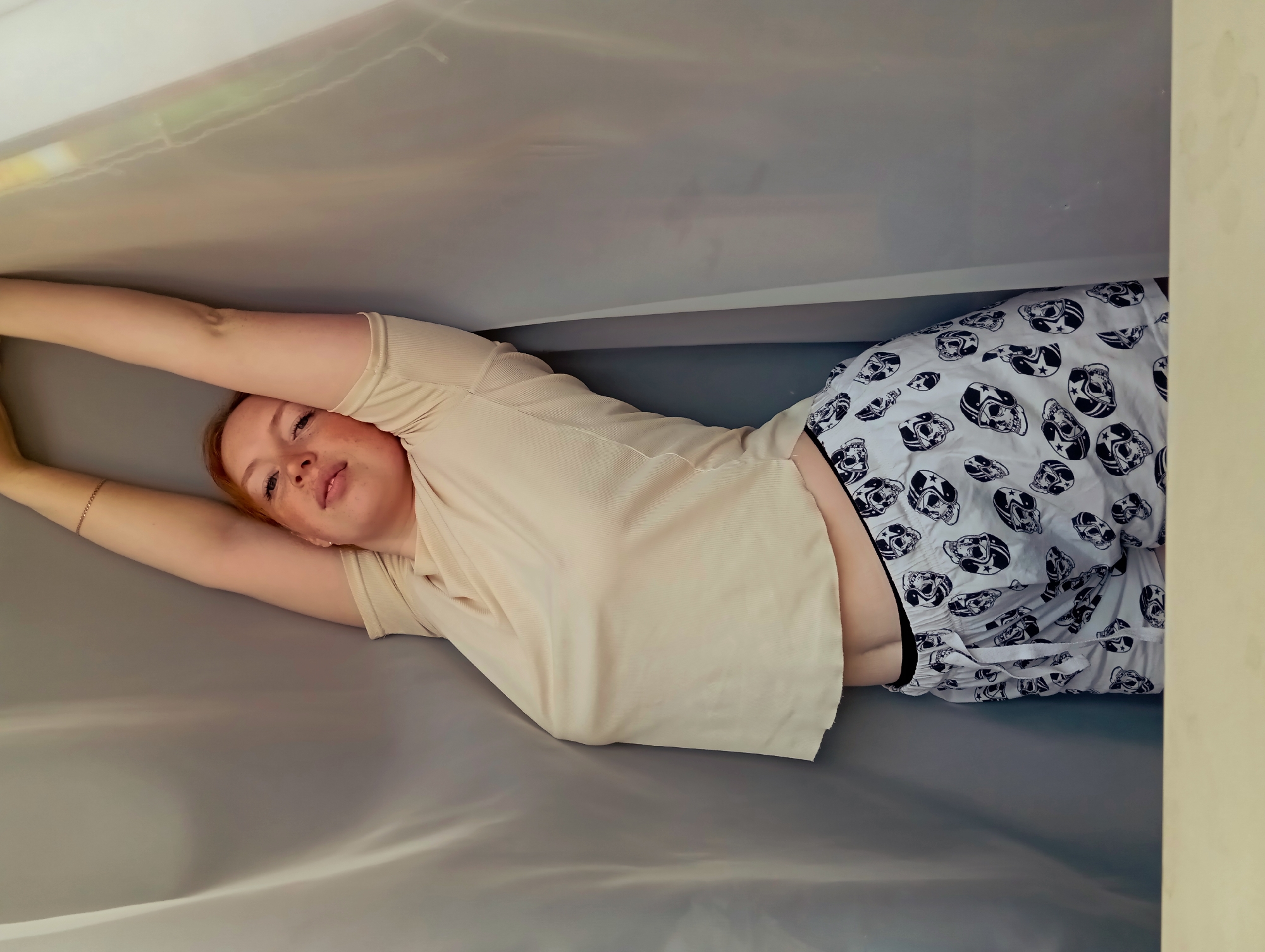 A young woman with reddish hair is lying down, apparently indoors. She is wearing a light tan t-shirt and patterned white and black shorts. Her arms are raised above her head, holding up a piece of light-colored fabric. The background and surrounding area are mostly in shades of grey and white. It is difficult to determine the location in Yurgamysh, Russia based on the provided image.