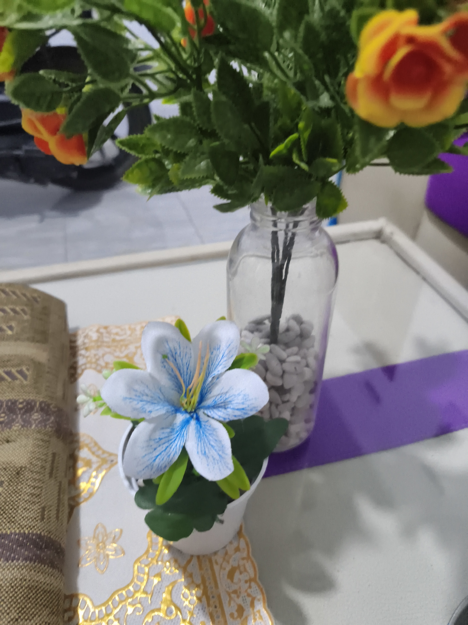 This is a close-up, eye-level, slightly out-of-focus photograph of artificial flowers on a table. In the foreground, a white pot contains a single light blue and white artificial flower with prominent blue veins. Behind it, a clear glass vase filled with small white pebbles holds a bouquet of artificial flowers with green leaves and orange and yellow blossoms. The table surface is covered with a decorative cloth that has a patterned border in gold and brown. A purple fabric band runs along the right side of the table. The background is blurred, showing what appears to be a dark object, possibly a vehicle, and a light-colored wall or surface. The lighting suggests an indoor setting with natural light. The image was taken in Pangkalpinang, Indonesia.