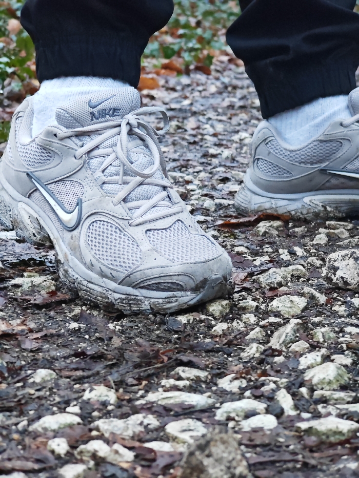The scene depicts the lower legs and feet of an individual standing on an unpaved, rocky, and muddy path, located in what is identified as Shumen, Bulgaria.
**Subject:** A person, visible only from the shins downwards.
**Apparel:** The individual wears black trousers or sweatpants, cuffed around the ankles, revealing white athletic socks.
**Footwear:** Two light grey athletic shoes, specifically Nike brand sneakers, are visible. The left shoe is prominent in the foreground, showing a mesh upper, synthetic overlays, white laces tied in a bow, a silver/white Nike "swoosh" logo on the side, and the "NIKE" text on the tongue. Both shoes exhibit significant accumulation of dark brown mud and dirt on their soles, midsoles, and lower uppers, indicating recent travel through wet or muddy terrain.
**Action:** The individual appears to be standing or momentarily paused while walking on the path, with the left foot slightly forward.
**Environment:** The ground is an uneven natural path composed of small, light-colored rocks, dark soil, and scattered wet, decaying brown leaves. The background is blurred, showing soft green and brown foliage, indicative of a wooded area or natural park setting. The overall conditions suggest a damp or recently wet environment.