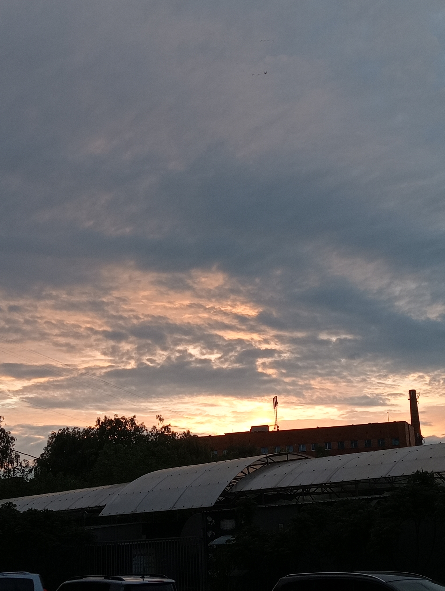 The image captures an urban sunset scene. The sky is predominantly cloudy with hues of orange and pink breaking through the cloud cover, suggesting the sun is setting. In the foreground, there is a structure with a long, sloping roof, likely a market or industrial building. Behind this structure, some trees and a building with a chimney and antenna on top can be seen silhouetted against the sunset. Several vehicles are parked in the foreground, partially visible. The location is likely a city, but the specific location remains unknown.