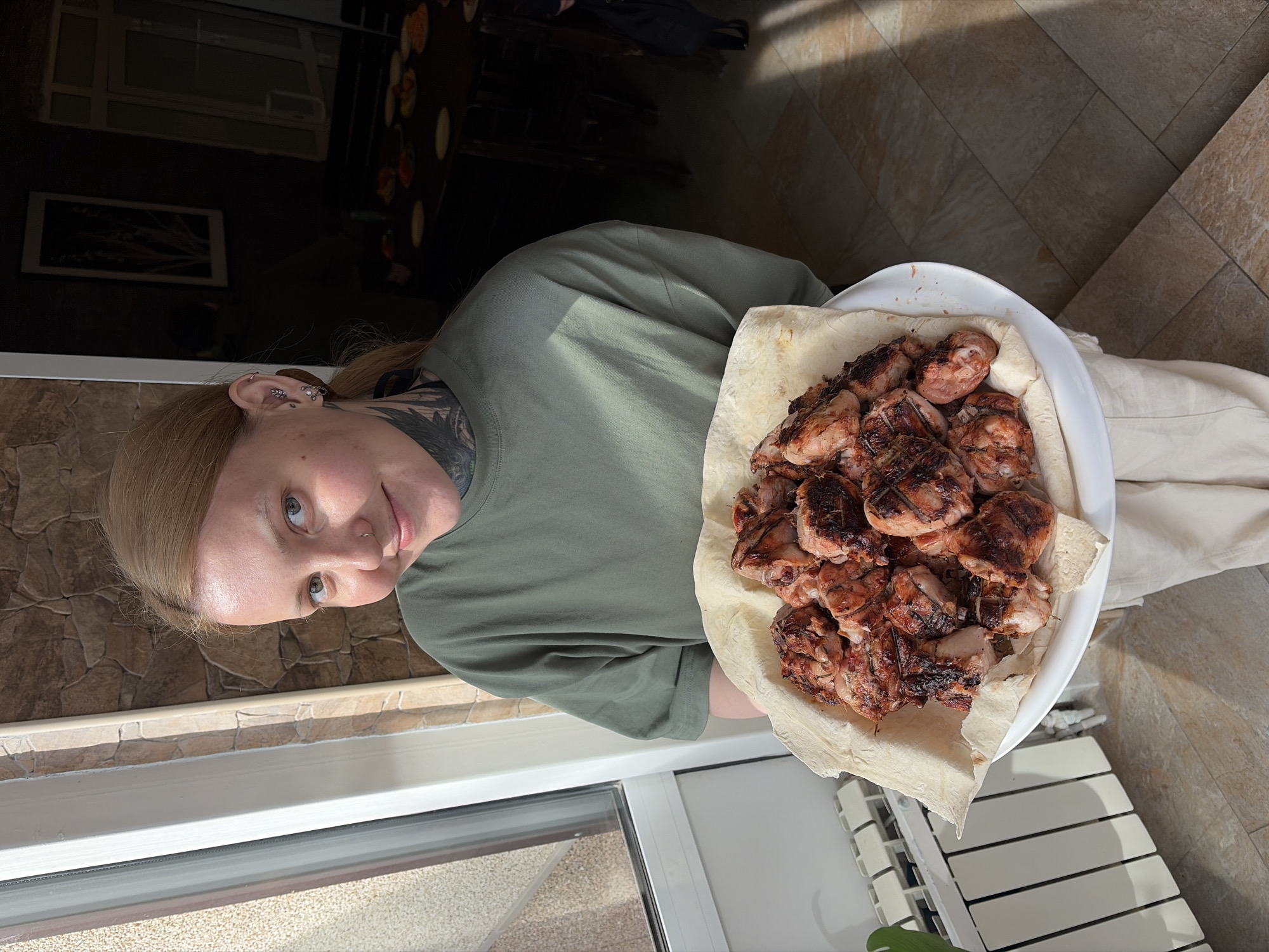 A woman with blonde hair, multiple ear piercings, and a neck tattoo is holding a white plate filled with cooked meat on top of a flatbread. The woman is wearing a green t-shirt and light-colored pants. The scene appears to be indoors, next to a window. The background includes a wall with a textured surface resembling stone, a radiator, and a glimpse of furniture in an adjacent room. While the location is specified as Chaikovsky, Russia, there are no visual elements in the image that definitively confirm this location.