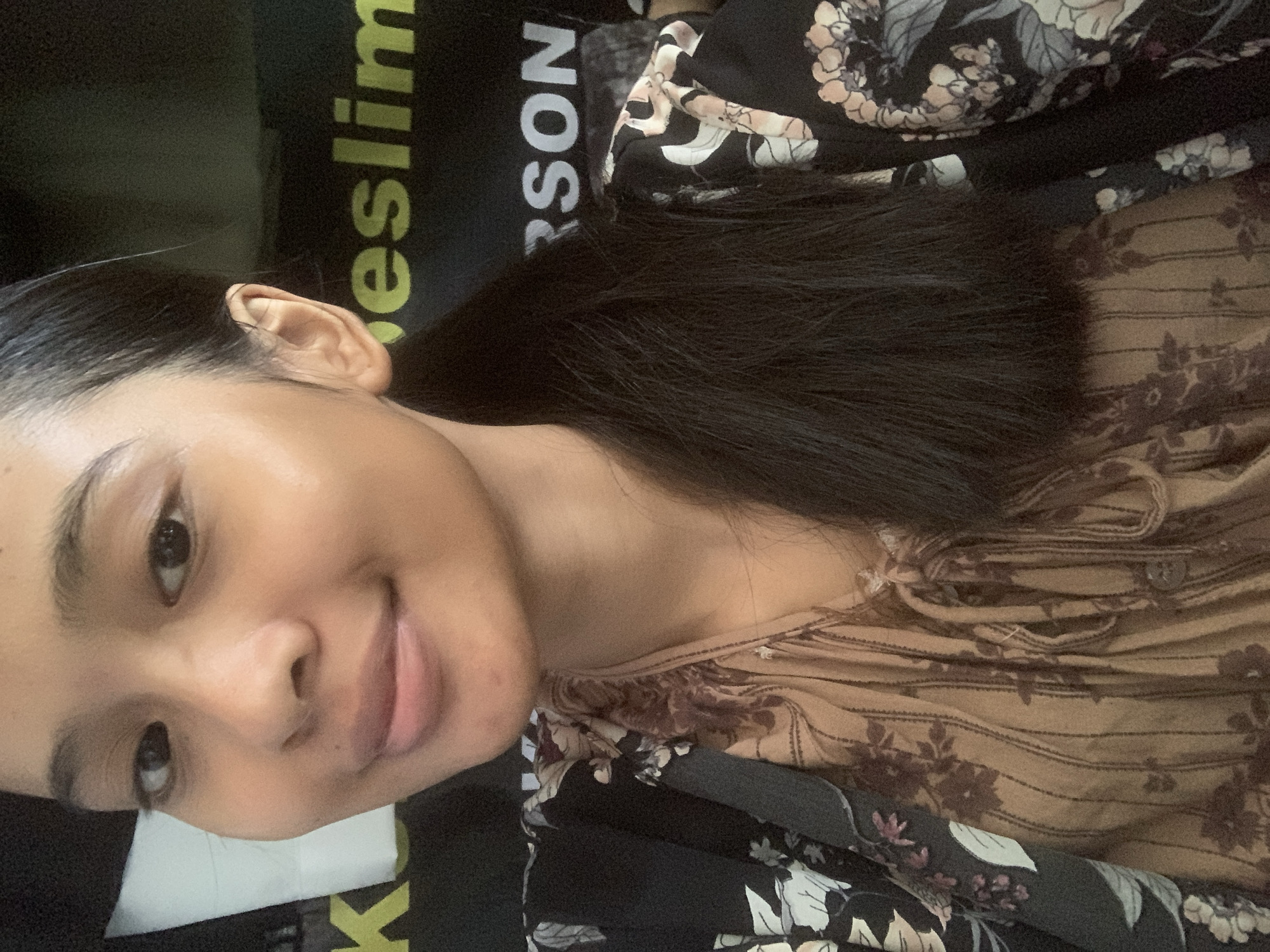 This is a close-up, medium shot of a young woman with dark hair pulled back into a ponytail. She is looking directly at the camera with a slight smile. The woman is wearing a floral print top. In the background, partially visible, are dark t-shirts with yellow and white lettering. The woman's left ear is visible, and her skin appears smooth and clear. The overall lighting suggests an indoor setting. There are no discernible elements that place this image specifically in Sungai Meriam, Indonesia, or any other specific city.