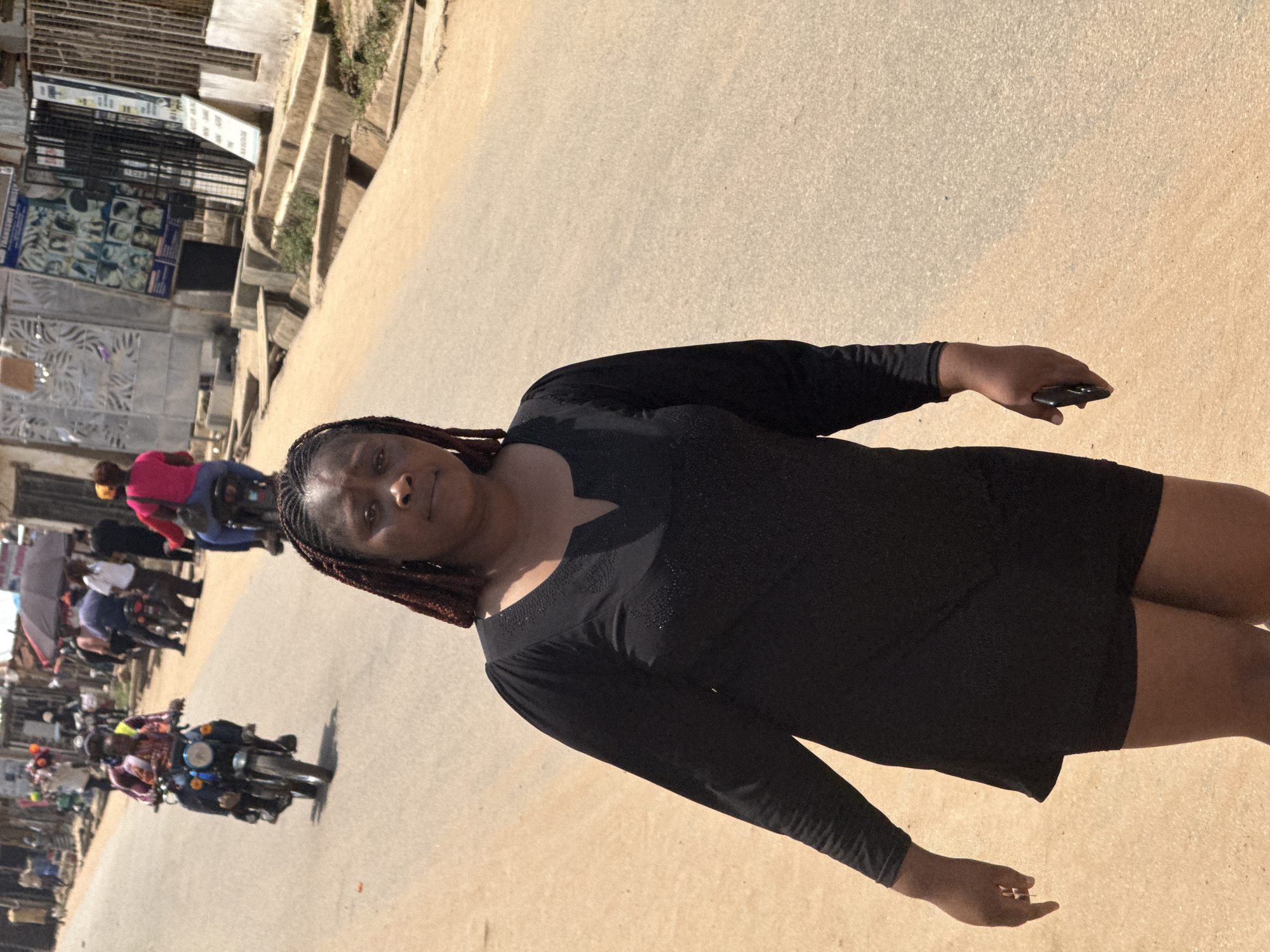 In Abuja, Nigeria, a woman with braided hair walks on a dirt road. She is wearing a black V-neck dress and black shorts. The road is dusty and appears to be in a neighborhood with shops and buildings on the side. In the background, there are several people on motorcycles, indicating a busy street. One person is riding a motorcycle in the foreground, facing left. Another person is on a motorcycle further in the background, also facing left. There are also pedestrians and stalls set up along the road. The overall scene is one of a bustling urban environment.