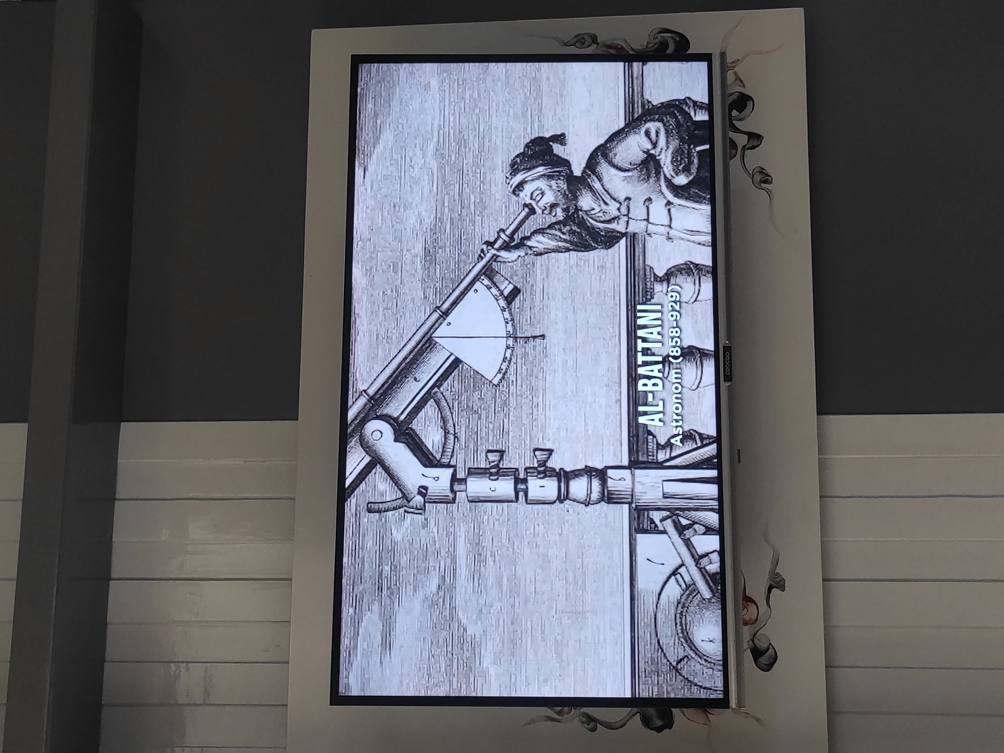 A digital display screen shows a black and white illustration of an astronomer, identified by text on the screen as "AL-BATTANI, Astronom (858-929)". The astronomer, wearing a turban and traditional clothing, is looking through a large, intricate astronomical instrument that appears to be a quadrant or astrolabe. The instrument has various arms, sighting devices, and a graduated arc. The background of the illustration is a stippled pattern, suggesting a sky or a textured surface. Below the illustration, the screen displays the text "AL-BATTANI" in a large, bold font, followed by "Astronom (858-929)" in a smaller font. There is a logo that reads "COOCAA" at the bottom of the screen. The display is mounted on a wall that has a decorative border with swirling, organic shapes. The wall itself is painted in alternating sections of dark grey and light grey vertical panels, with a darker horizontal band across the top. The display and wall decoration are positioned in front of a background that consists of vertical light grey panels. This scene is located in the city of Sumedang, Indonesia.