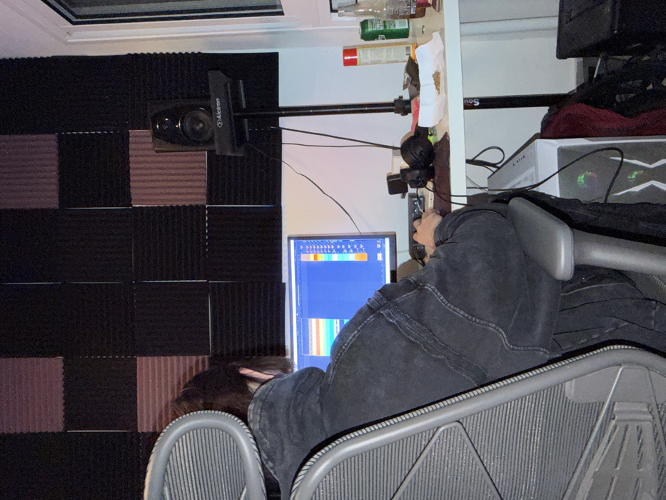 An individual is seated in a grey ergonomic office chair, facing away from the camera, engaging with a computer setup within a room in Migdal HaEmeq, Israel. The person wears a dark, possibly black, denim jacket or heavy hoodie, with short dark hair visible. Their hands are positioned over a keyboard and mouse on a white desk.
The computer monitor displays a Digital Audio Workstation (DAW) software interface featuring multiple colored horizontal tracks. To the right of the monitor, on the desk, a black studio monitor speaker labeled "Alctron" is mounted on a stand. Various items are present on the white desk, including: a brown glass bottle, a green aluminum can, a red aerosol can, a pair of black headphones, and a small black electronic device. Notably, a small pile of green, leafy, granular substance rests on a crumpled piece of white paper or tissue. A white PC tower with illuminated internal components is visible on the floor beneath the desk.
The wall behind the computer setup is covered with sound-absorbing acoustic foam panels, arranged in a checkerboard pattern of dark grey/black and reddish-brown or dark purple sections. A white wall and a window frame are visible on the right side of the image. Multiple black cables are connected to the electronic equipment.