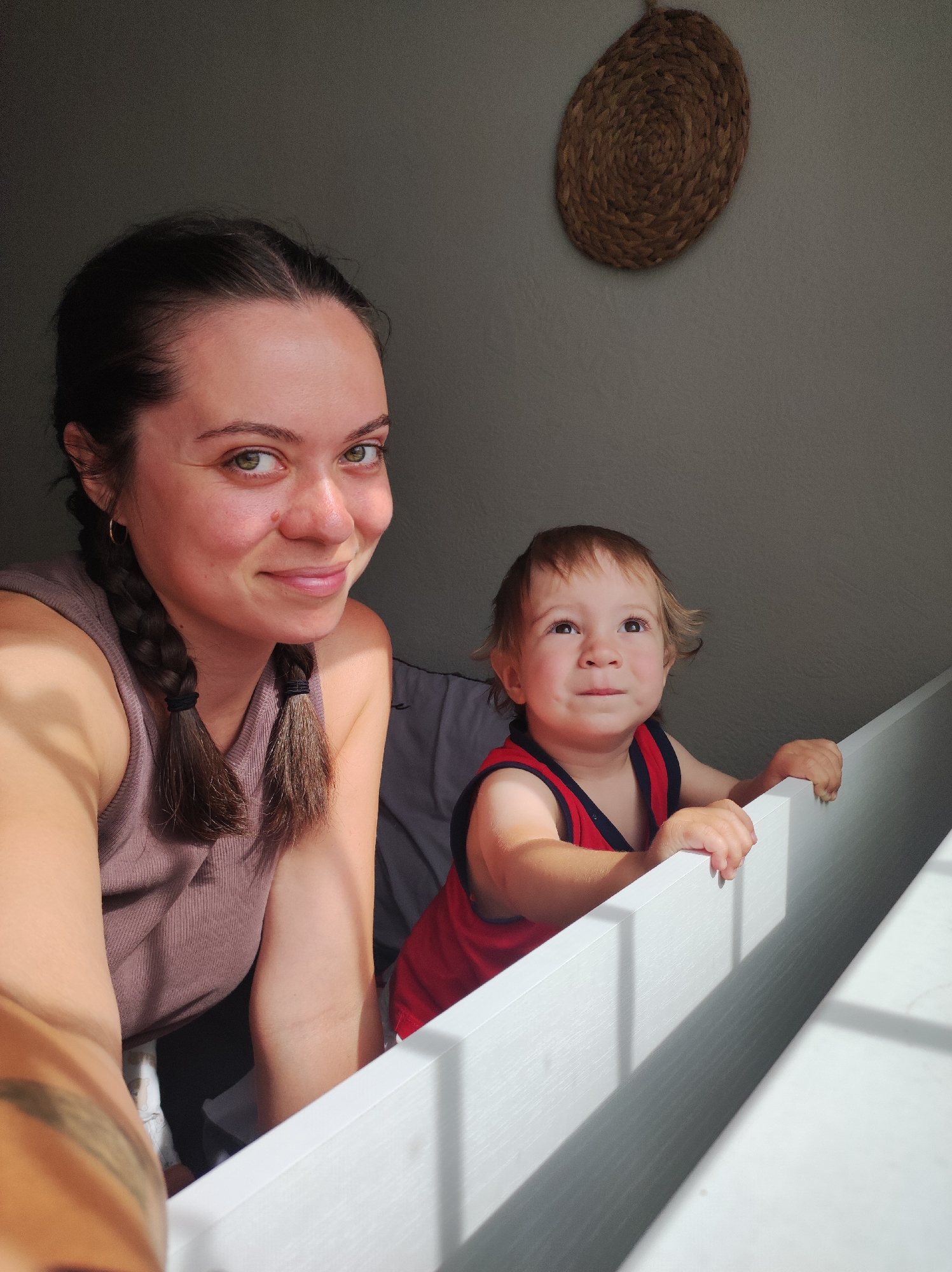 This media file is a photograph featuring a woman and a young child. The woman, in the foreground, has dark hair styled in two braids and is wearing a taupe tank top. She is smiling and looking towards the camera. To her right and slightly behind her is a young child with light brown hair, wearing a red and blue tank top. The child is looking upwards and to the right, with hands resting on a white railing. The background is a muted gray wall with a woven circular decoration hanging on it. Light is streaming in from the right, casting shadows on the white railing and the subjects. The overall mood appears warm and intimate, suggesting a casual moment captured at home. The time of day is likely daytime due to the strong sunlight and shadows. The location context is Chaikovsky, Russia, though this information is not visually apparent in the image itself. There is no visible text in the image.