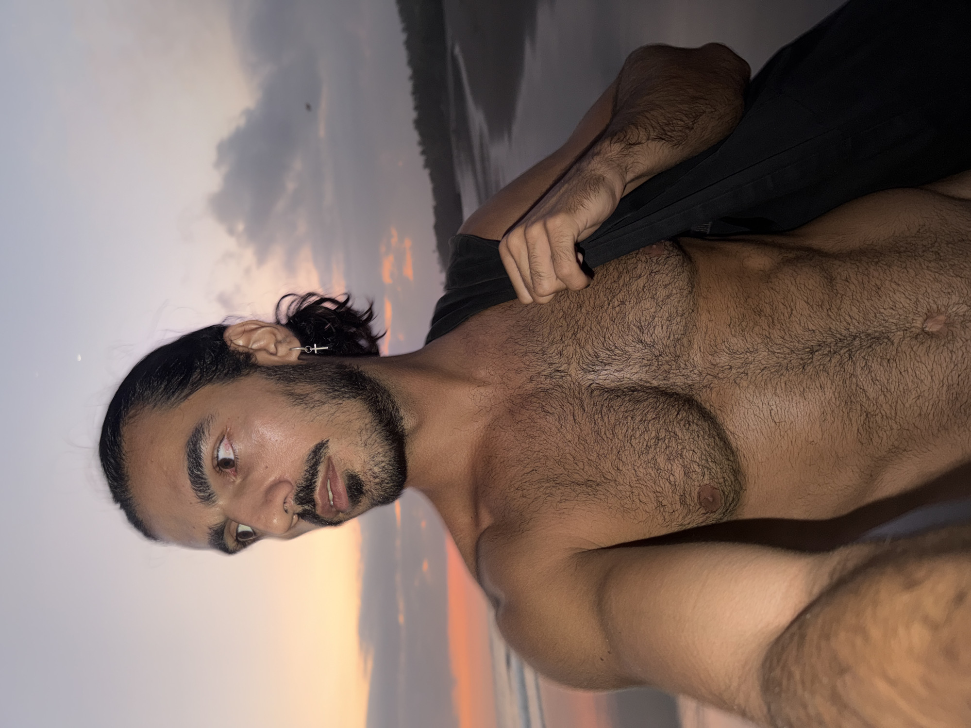 A shirtless man with dark hair, a full beard, and a mustache is depicted in a medium-shot, facing partially right and looking left with a serious expression. His dark hair is pulled back into a bun or ponytail. He wears a silver-colored hoop earring with a small cross pendant in his visible right ear. His torso is muscular and covered in dark body hair. A black strap, possibly from a bag, is slung over his right shoulder and held by his right hand. The background reveals a coastal scene at either sunrise or sunset, consistent with a beach location in Selemadeg, Indonesia. The sky displays a gradient of warm orange and pink hues near the horizon on the left, transitioning to cooler purples and greys with scattered clouds towards the right. A faint crescent moon is visible in the upper right sky. Gentle waves are seen on the ocean to the left, and a sandy beach curves along the distant shoreline to the right, bordered by dark foliage.