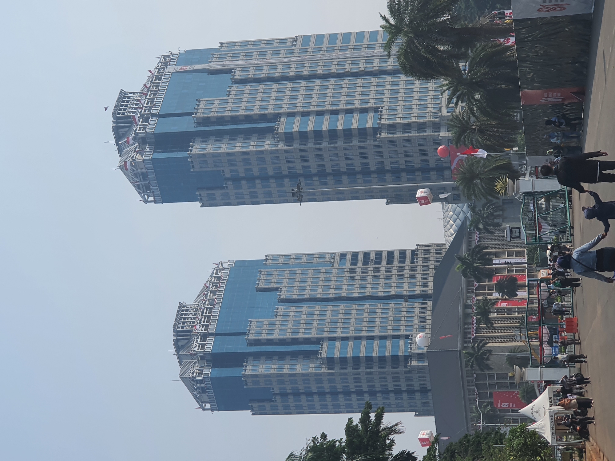 In this medium-angle, eye-level shot of the Istiqlal Mosque complex in Central Jakarta, Indonesia, two towering blue-glass skyscrapers dominate the skyline. The buildings are illuminated by natural daylight under a hazy, pale sky. In the foreground, a paved walkway leads towards a crowd of people and a stately building with a dark roof, which is likely part of the mosque complex. Palm trees are scattered throughout the scene, some in the foreground and others behind the buildings. Red and white banners adorn the front of the building and are visible on some of the skyscrapers. A few people are walking on the walkway, some are in small groups, while others are walking individually. The overall scene depicts a public gathering or event in an urban setting, likely a tourist destination or a place of cultural significance.