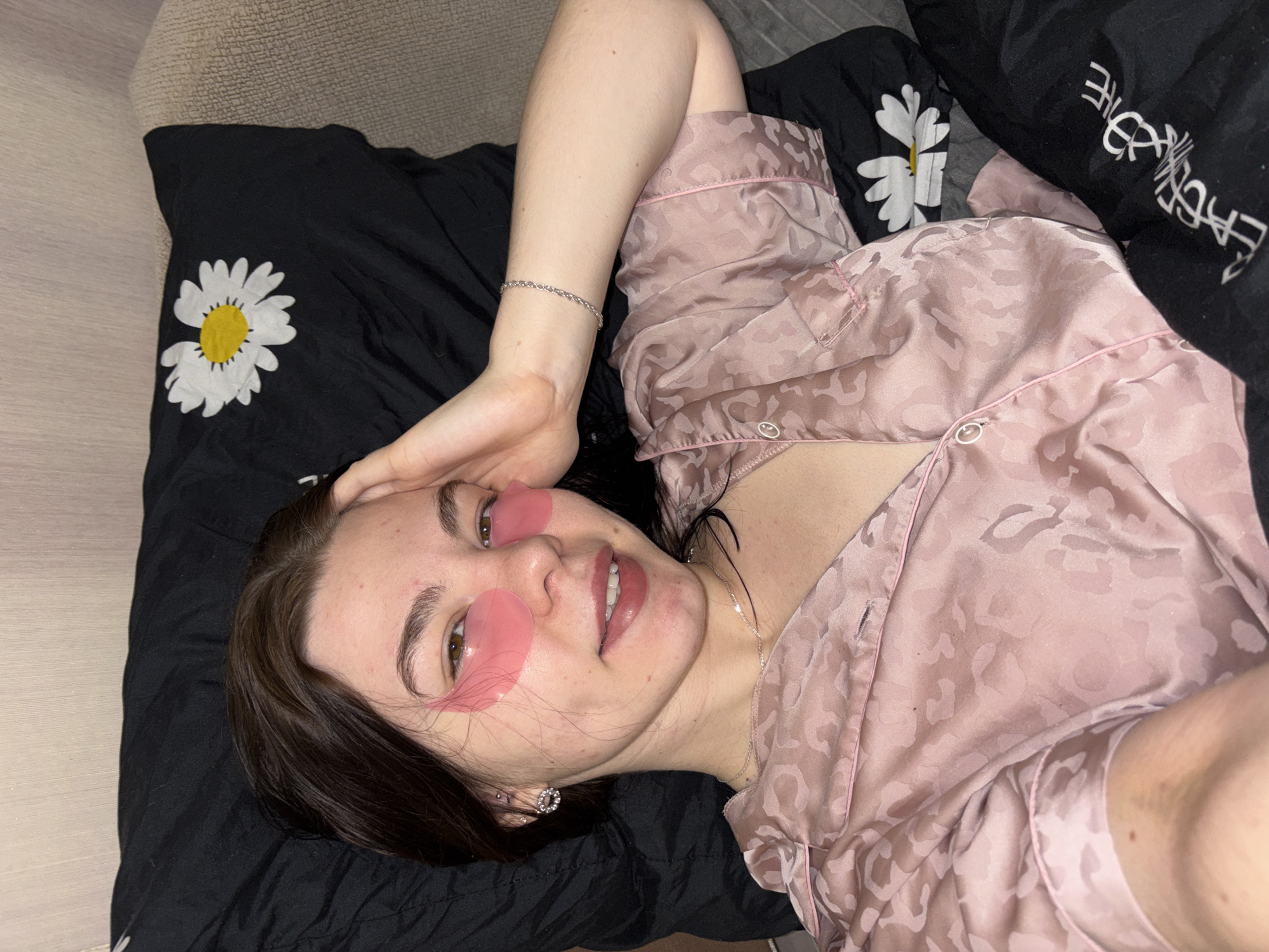 A woman is lying down, possibly on a bed, as indicated by the pillows and sheets. She is wearing a pink patterned pajama top and pink under-eye patches. Her hair is dark. She is wearing jewelry including a bracelet, earrings, and a necklace. The sheets are black with a white daisy design. The location is in Perm, Russia.