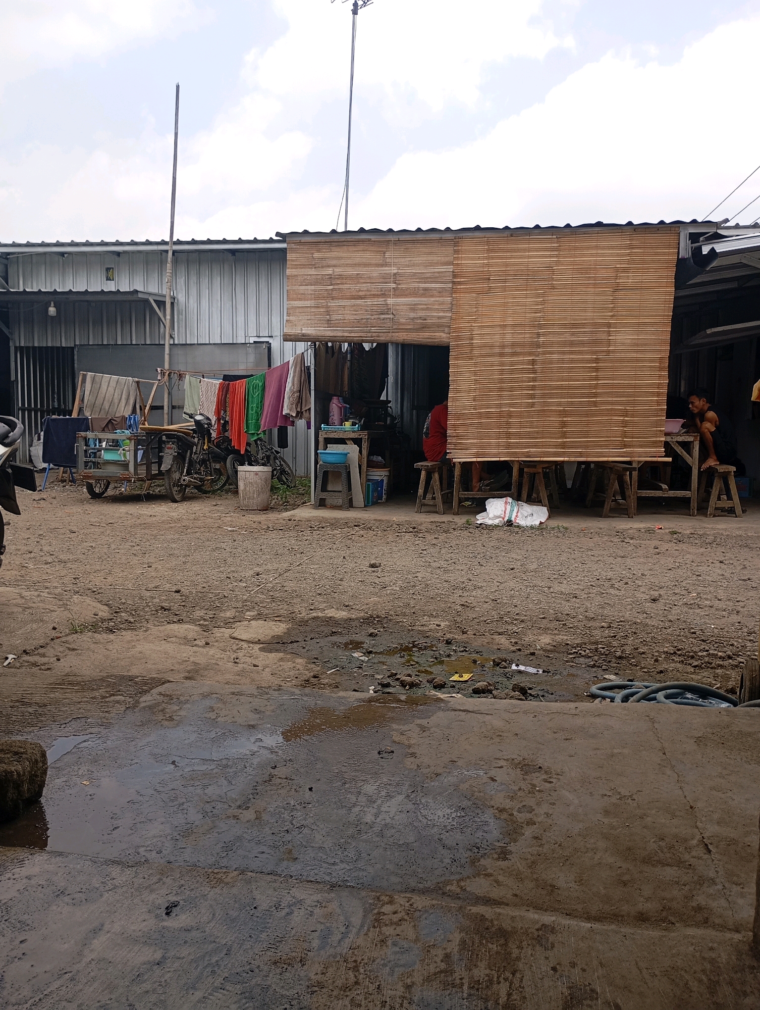 The image depicts a scene in Tasikmalaya, Indonesia, likely in a semi-urban or rural area, characterized by informal structures and a dusty, unpaved ground. In the foreground, a wet concrete area with puddles suggests recent rain or water spillage, with debris scattered around. A coiled blue hose is visible to the right.
Further back, the ground is rough and uneven, with scattered rocks and smaller debris. A small puddle with some yellow plastic is present. Clothes are hanging on a line strung between two poles, with various garments in different colors, including red, green, and pink. Adjacent to this is a small cart with two wheels, a motorcycle, and a cylindrical trash bin.
A simple structure with a roof made of corrugated metal and walls possibly constructed from bamboo or reeds forms the background. A large section of this structure is covered by a bamboo matting. Underneath this covered area, several wooden stools are placed around a table-like structure. Two individuals are visible: one person in a red garment is partially obscured behind the bamboo matting, and another person in a dark shirt is sitting on a stool to the right, looking towards the left. A white plastic bag lies on the ground under the table. Another smaller structure with metal siding is located to the left of the main building. A single bare lightbulb hangs from the metal structure. The sky above is cloudy.