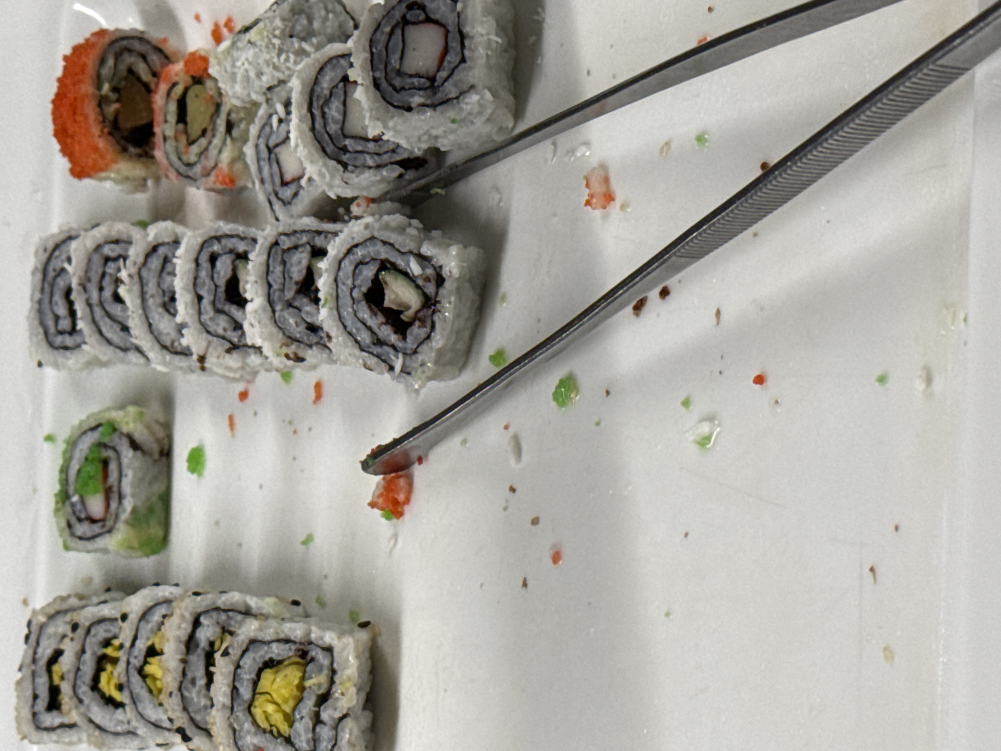 The image displays a tray of sushi rolls, likely at a buffet or restaurant, in Nordhausen, Germany. The sushi is arranged in rows on a white surface, with some scattered pieces of green and orange roe. A pair of metal tweezers is positioned in the foreground, resting on the white surface. The sushi rolls themselves vary in appearance; some are coated with white sesame seeds and black sesame seeds, while others have a green exterior, possibly avocado. Several rolls appear to contain crab stick and cucumber. The overall scene suggests a casual dining environment where sushi is served.