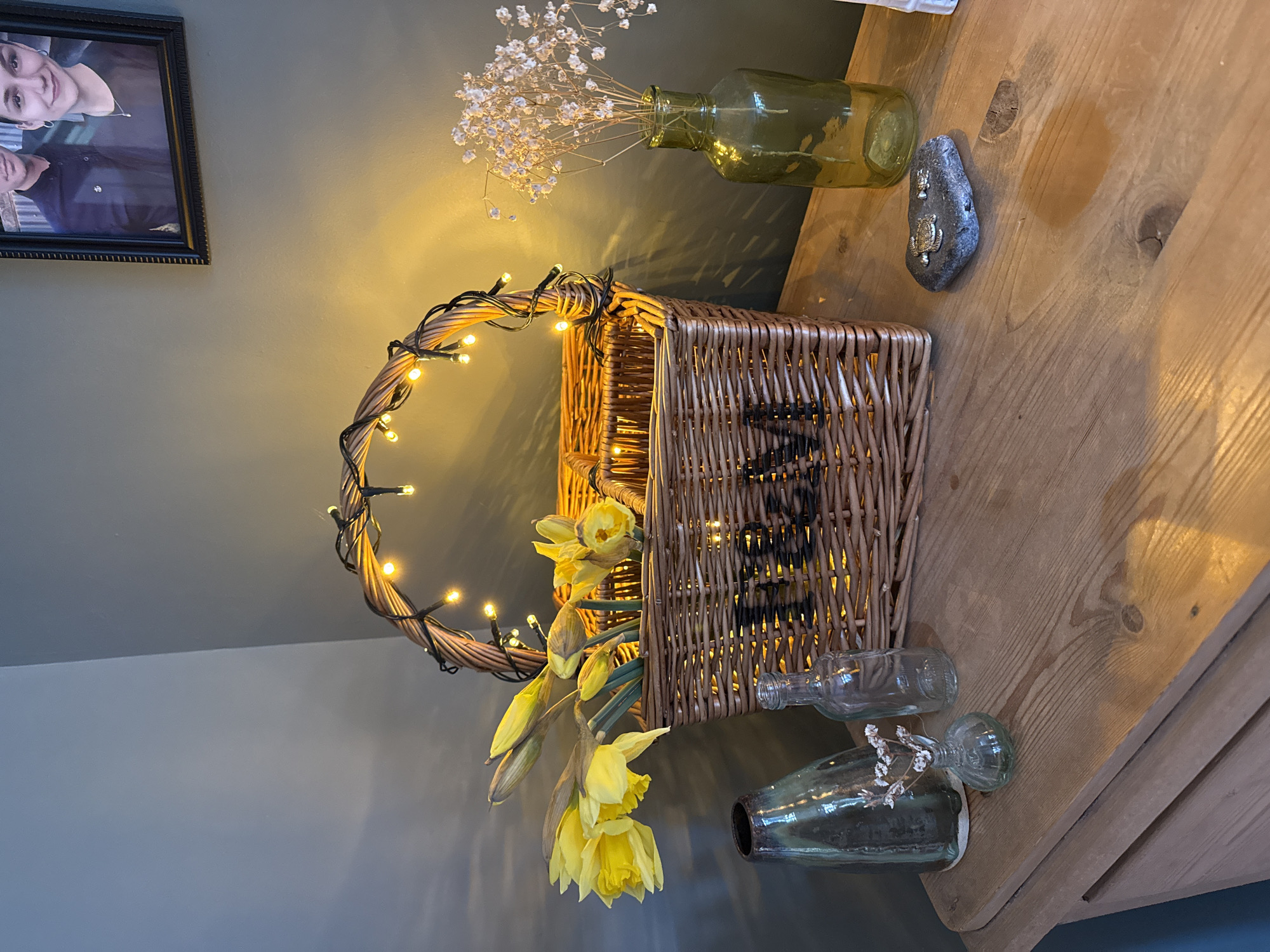 A still life scene is depicted indoors, with a wooden dresser surface serving as a display for various objects. A wicker basket, embellished with small yellow fairy lights around its handle, is the focal point. Daffodils are arranged within the basket. To the right of the basket are two small glass bottles. Behind the basket is a larger green glass bottle holding baby's breath flowers. A grey stone with inlaid turtle decorations is positioned near the larger bottle. A framed photo hangs on the wall to the left. The wall is painted a light grey colour. Given the location context provided (East Suffolk, United Kingdom), the scene likely represents a domestic interior.
