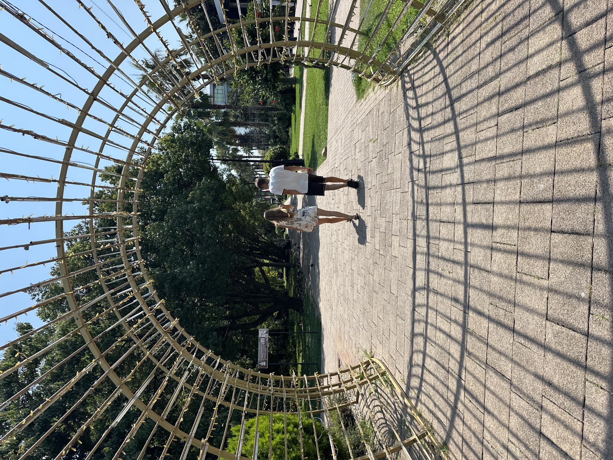 The image depicts a park scene in Sochi, Russia, on a sunny day. A couple, a man and a woman, walk away from the camera under a large, open archway constructed of metal tubing and decorated with small lights. The archway casts a grid-like shadow on the paved walkway made of interlocking stone tiles. Lush green trees and grass surround the walkway, with palm trees visible in the background. The sky is clear and blue.