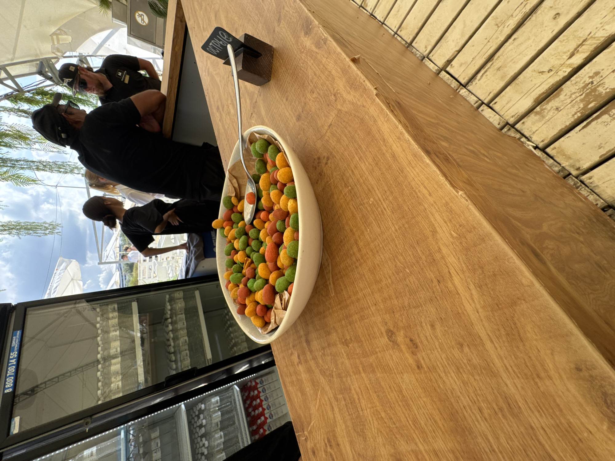 In Vityazevo, Russia, at what appears to be an outdoor bar or food stall, a white bowl filled with multi-colored, coated peanuts sits on a wooden counter. A silver spoon rests in the bowl. To the right of the bowl, a small dark wooden stand holds a black sign with white Cyrillic text, likely indicating the product. Behind the counter, several people are visible, including two men wearing black t-shirts and baseball caps, and two women in the background. One woman, wearing a black t-shirt, stands with her hands behind her back. To the left of the counter, a large glass-fronted refrigerator is filled with bottled water. The scene is outdoors, with a cloudy sky and foliage visible in the upper portion of the image, suggesting a warm, possibly summer day. The wooden counter and surrounding deck indicate a casual, possibly recreational setting.