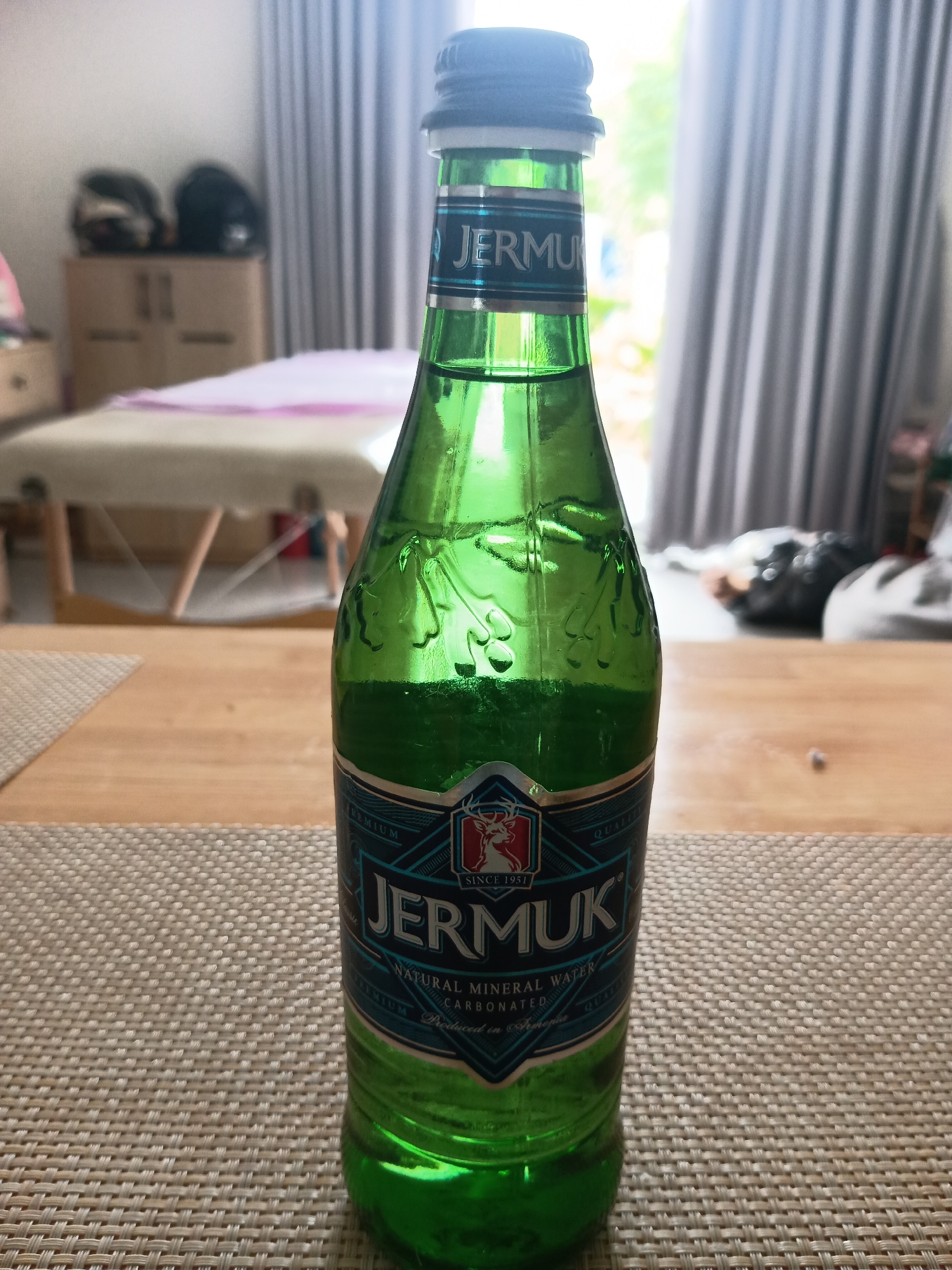 A green glass bottle of Jermuk Natural Mineral Water stands centrally on a light wooden table covered by a light-brown woven placemat. The bottle features a dark blue/black screw cap and a prominent label with "JERMUK" in white lettering on a blue background, adorned with silver borders and a red deer head logo. Text visible on the label includes "SINCE 1951", "NATURAL MINERAL WATER", and "CARBONATED", with "PREMIUM QUALITY" also partially visible.
The background is softly blurred, revealing a brightly lit indoor space, consistent with a daytime setting. Light grey vertical curtains hang in the background, suggesting a window or doorway, through which bright natural light enters. To the left, a light-colored wooden cabinet topped with two dark motorcycle helmets is visible. In the middle distance, a light-colored treatment or massage table with a pink covering is present, indicating a potential spa, clinic, or personal care room. The scene is devoid of people and conveys a tranquil, clean environment in Mũi Né, Vietnam.