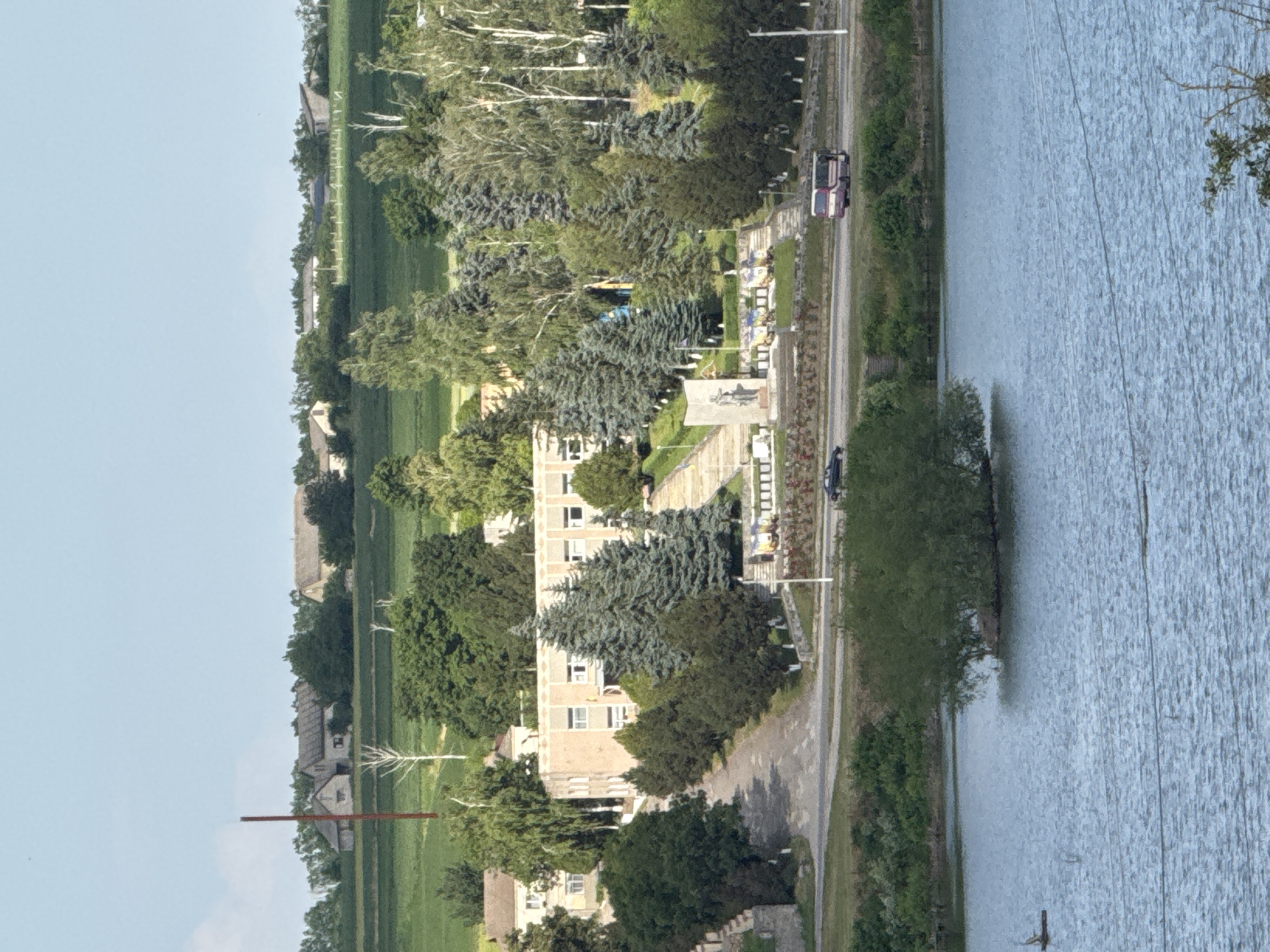 The image shows a wide view of a rural or suburban landscape dominated by a body of water in the foreground and a building complex on a hill in the midground. The water, appearing to be a lake or large pond, fills the lower portion of the frame and has a slightly rippled surface under a bright sky. A small, tree-covered island is visible in the water.
On the hill, a long, multi-story building with a light-colored facade and visible windows is the most prominent structure. It is surrounded by a variety of trees, including tall, dark green coniferous trees and lighter, more deciduous trees. A paved road leads up to the building, and a set of stairs with a monument at the top is situated in front of it. Several other buildings, appearing to be smaller and residential or agricultural in nature, are scattered in the background on rolling hills covered in green fields. A tall, thin, reddish-brown structure, possibly a radio tower or a chimney, is also visible on the distant horizon.
A white and purple bus is driving on a road in front of the building complex, and a dark-colored car is visible further down the road. The environment appears to be a sunny day with a clear sky. The overall impression is one of a tranquil, possibly historical, setting in a non-urban area.