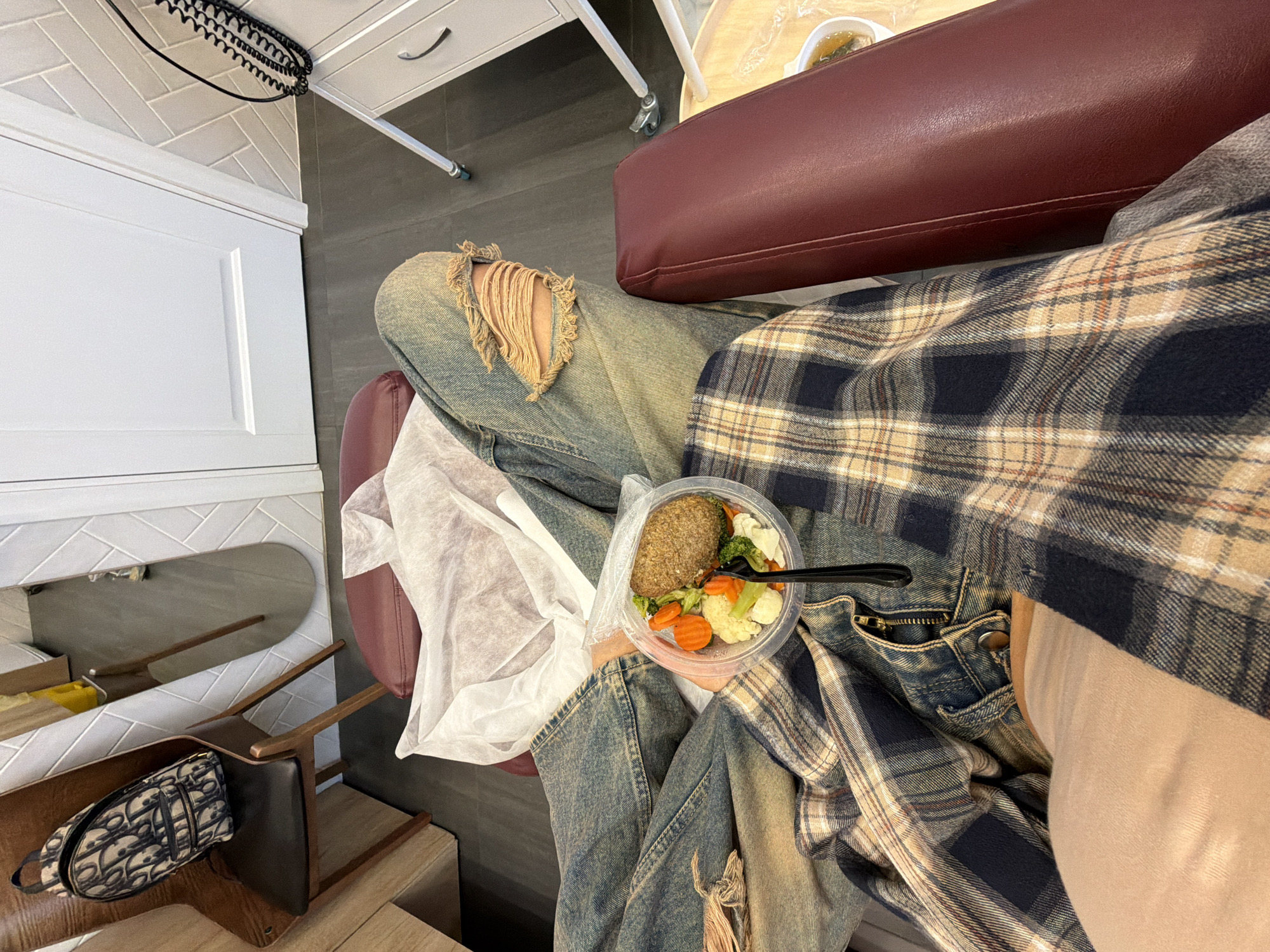 An elevated, slightly oblique shot captures a person seated indoors, likely in Krasnoyarsk, Russia, based on context clues. The individual is wearing ripped blue jeans and a tan and navy plaid shirt. A clear plastic container of food rests on their lap, containing what appears to be a large meatball or patty surrounded by steamed vegetables such as carrots, broccoli, and cauliflower. A black plastic fork is stuck in the food. To the left of the person's lap is a white plastic bag. The person is seated in a red chair, and a purse with a pattern is on a wooden chair underneath a mirror. Behind the chair is a white cabinet with a metal handle.