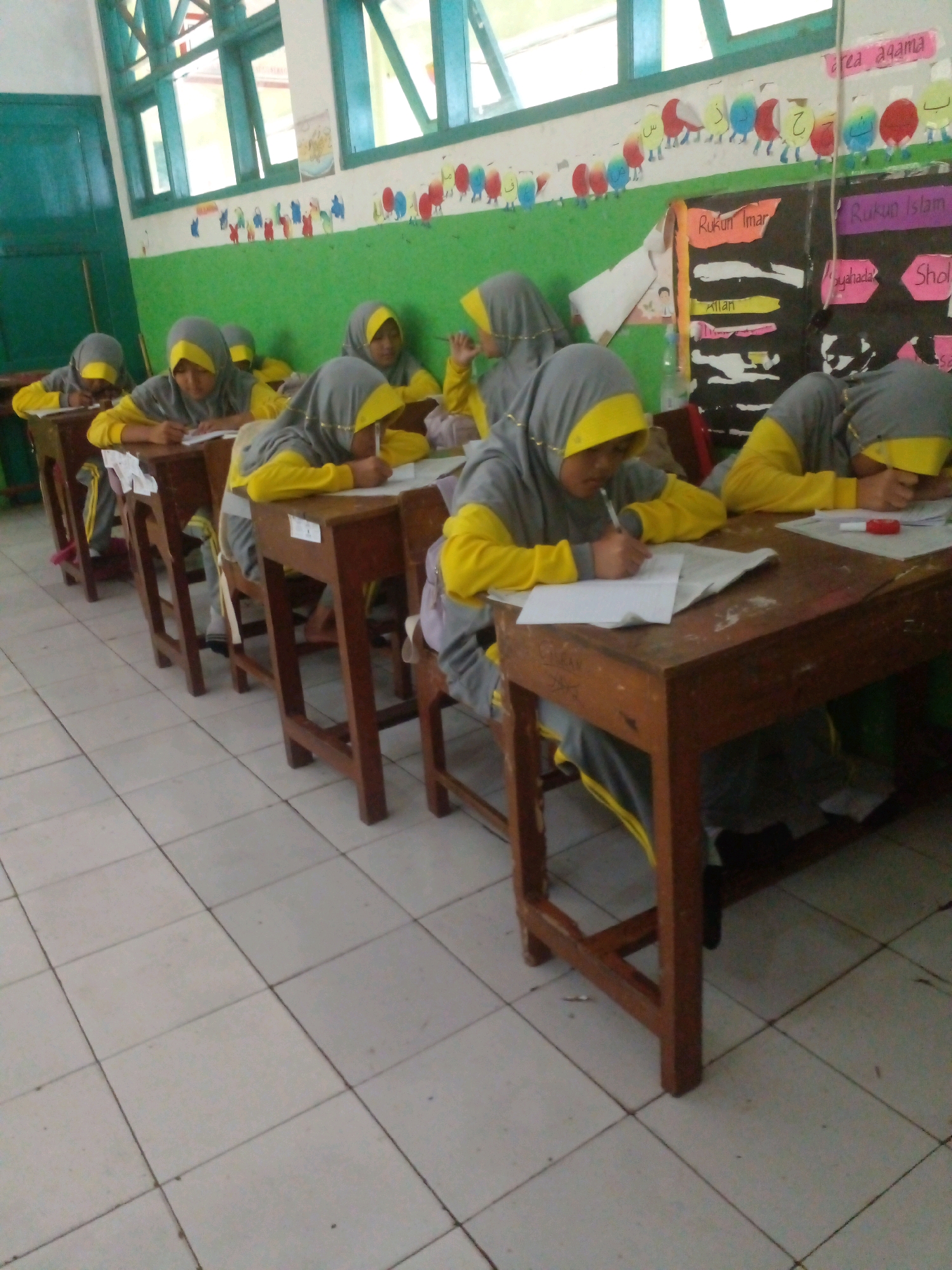 The image depicts a classroom scene in Banyuputih, Indonesia, likely a school for young girls. Several students, all wearing grey hijabs with yellow trim and matching yellow and grey uniforms, are seated at wooden desks, engaged in writing. The desks are arranged in rows, and the floor is tiled with white squares. In the background, there are colorful decorations on the wall, including what appears to be an alphabet chart with cartoonish figures and Arabic lettering. One of the students in the foreground is clearly visible writing in a notebook. Another student further back is resting her head on her desk, possibly fatigued. The overall atmosphere suggests a typical school day in progress.