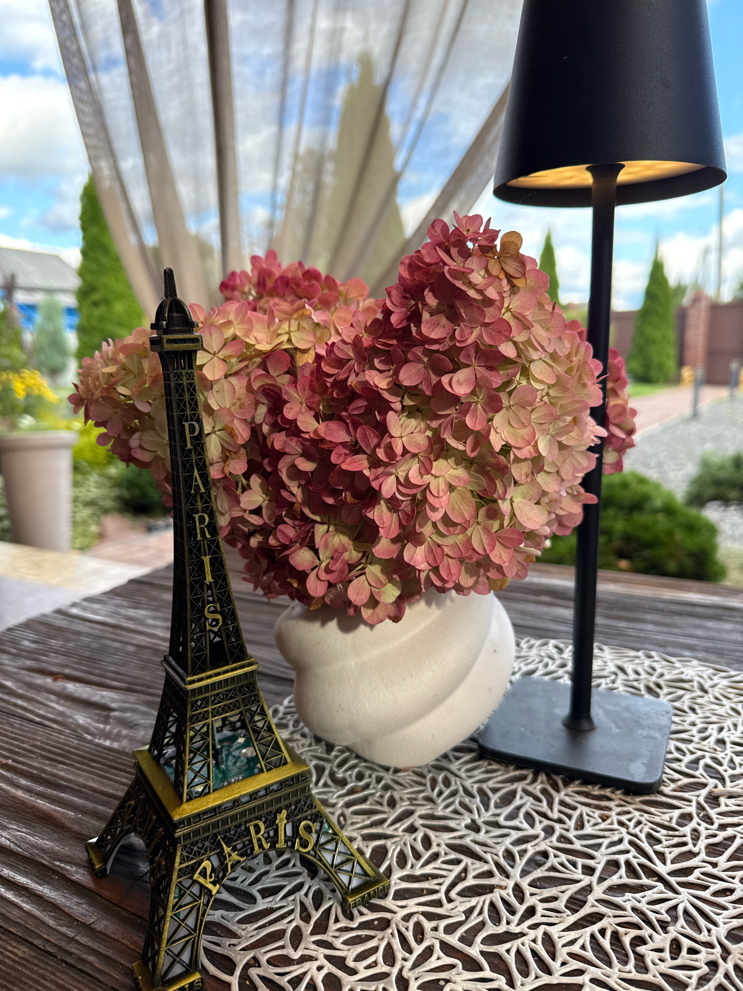 A miniature Eiffel Tower replica stands on a wooden surface next to a white vase containing dried hydrangeas. The Eiffel Tower is detailed with the word "PARIS" inscribed on its base. Behind the vase and Eiffel Tower, a black table lamp with an illuminated shade is positioned. The objects are placed on a decorative white placemat with an intricate leaf pattern. In the background, sheer curtains hang, revealing a glimpse of an outdoor scene with trees, a building, and a cloudy sky. The location is identified as being in the city of Slawharad, Belarus.