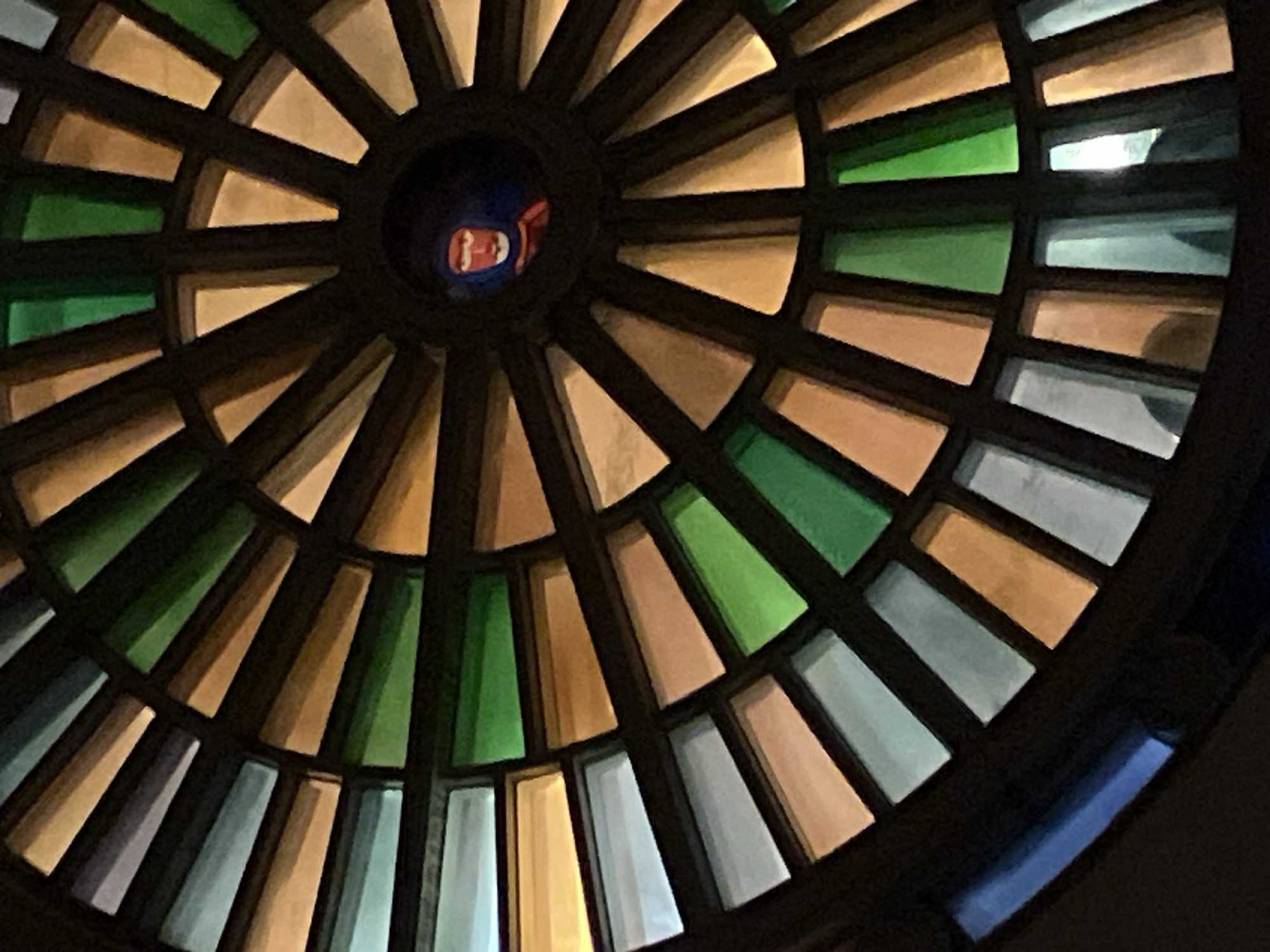 The image shows a circular stained glass ceiling or window, composed of numerous trapezoidal panes of various colors including green, tan, blue, and white. The panes are arranged in a radial pattern with dark ribs separating them. In the center of the circle, a face, possibly a reflection, is visible. The location is Unknown, Unknown.