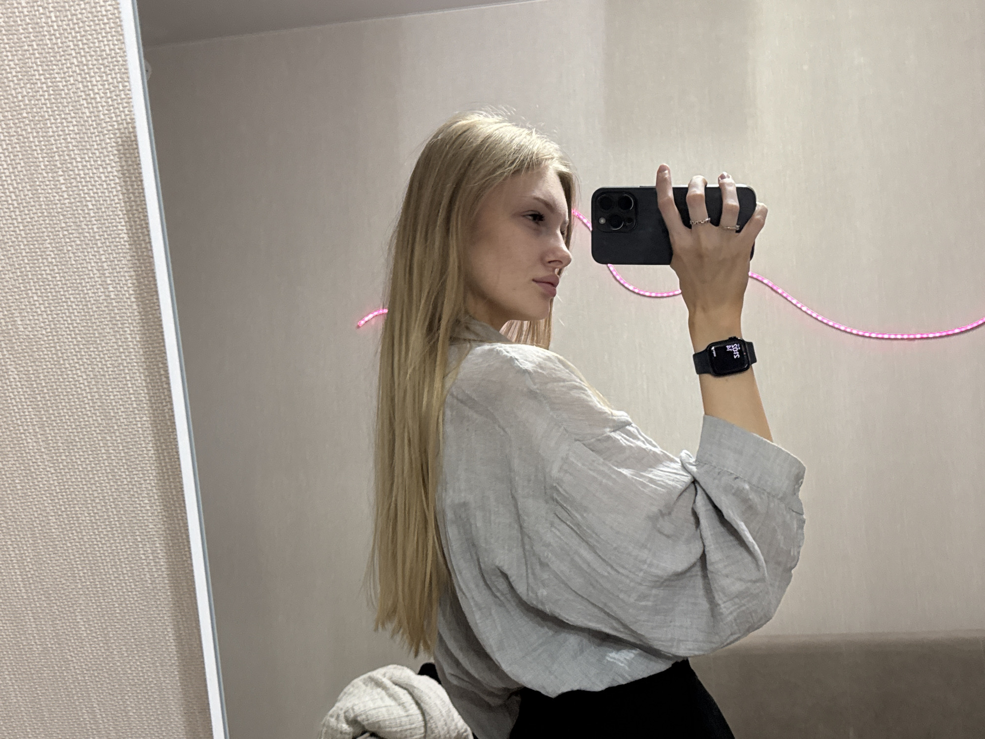 A young blonde woman with long hair takes a mirror selfie in what appears to be a residential interior, possibly a bedroom, in Perm, Russia. She is wearing a light gray, oversized, textured shirt and a black skirt. On her left wrist, she wears a black smartwatch. Her right hand is holding a black smartphone, taking the picture. She is wearing two silver rings on her right hand. A pink LED strip light is visible in the background, forming a circular shape on the wall. The background wall is a neutral light color, and the left side of the image shows a textured beige wall, possibly a headboard or a textured partition. The lighting appears to be artificial.