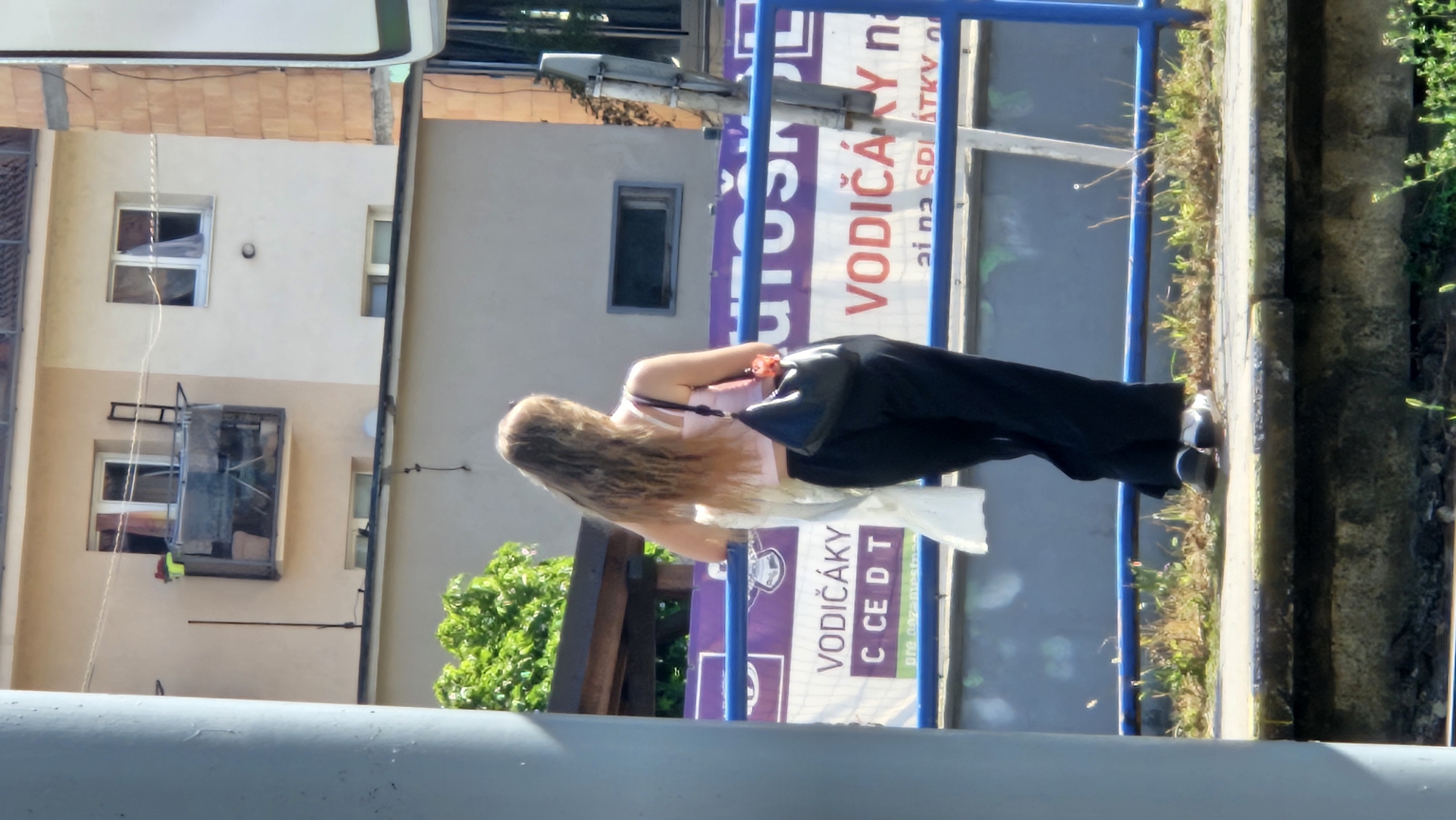 A young woman with long, wavy blonde hair is standing on a train platform in Prešov, Slovakia. She is facing away from the camera, looking towards a building in the background. She is wearing a pink crop top and black wide-leg pants. A black purse is slung over her shoulder, and she is holding a white garment in her left arm. A blue railing runs along the platform in front of her, and a banner with Slovak text is visible behind it. The building behind her is a multi-story structure with balconies and windows. The platform is elevated and appears to be made of concrete. There is some greenery and weeds growing between the platform and the track area. The overall lighting suggests it is a sunny day.