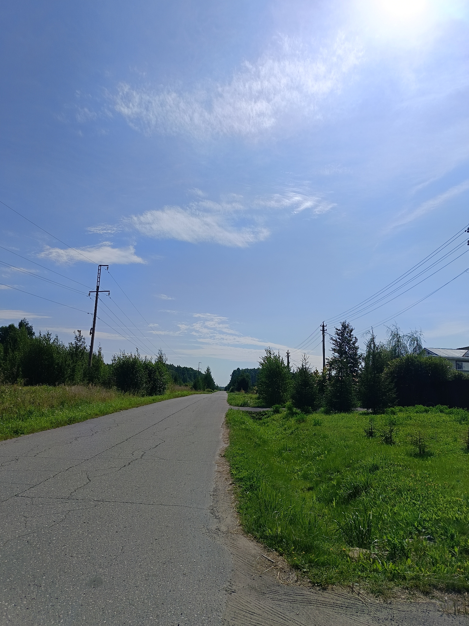 An asphalt road stretches into the distance, flanked by lush green grass and dense trees on either side. Power lines supported by tall wooden poles run parallel to the road, disappearing into the horizon. The sky above is a vibrant blue, dotted with wispy white clouds. The sun is shining brightly, casting a warm glow over the scene. In the distance, a house with a white roof can be seen peeking through the trees. The road surface is cracked, suggesting age and wear. The overall impression is one of a rural landscape on a clear, sunny day.
