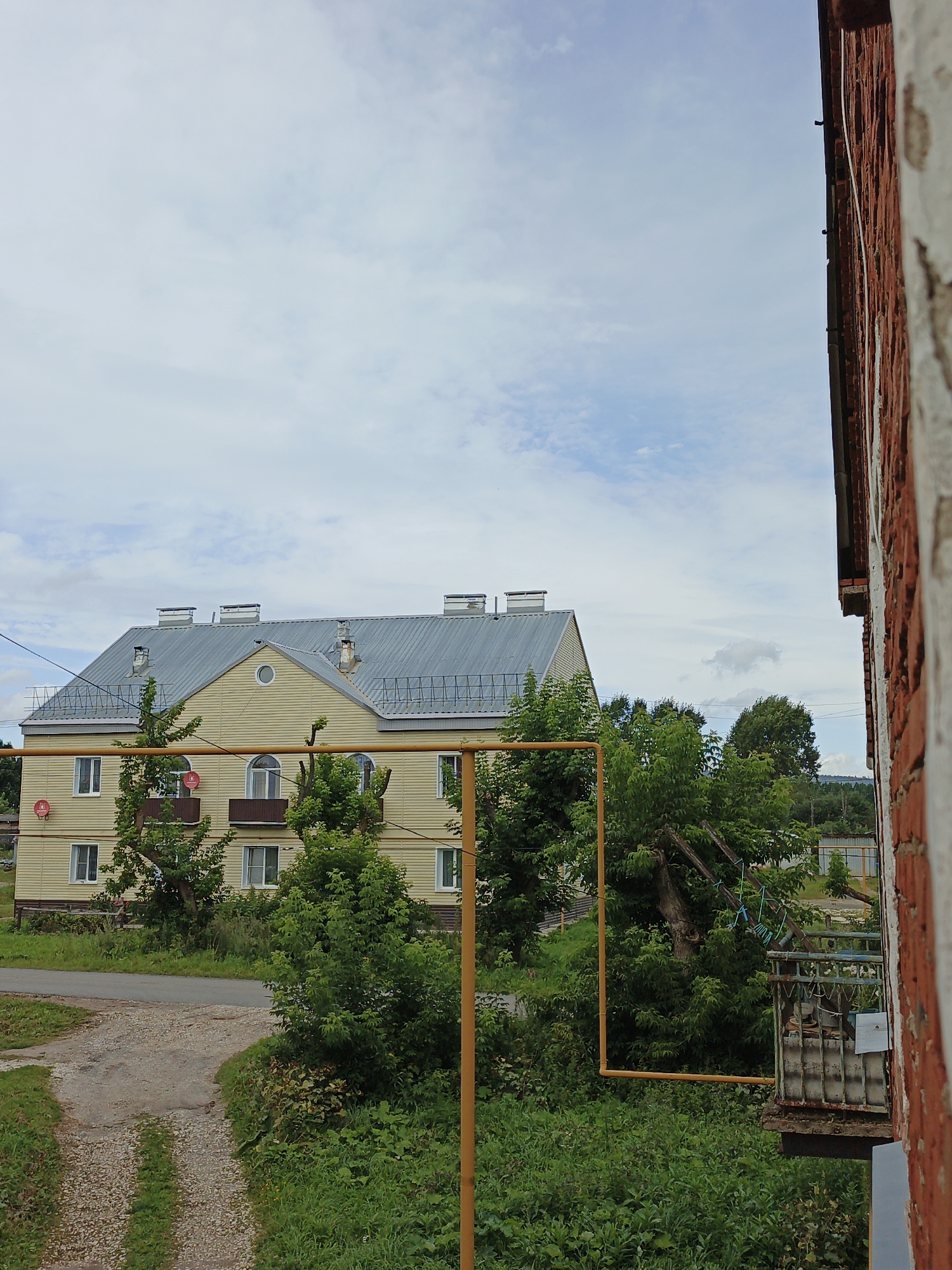 The image captures a view of a pale yellow, two-story apartment building with a corrugated metal roof, situated in Medvedyovka, Russia. Several dormer windows with chimneys are visible on the roof. The building has multiple windows and two small balconies with red satellite dishes mounted on the exterior wall. A yellow pipe runs horizontally in front of the building, partially obscured by green trees. The ground in front of the building is a gravel path that curves towards a paved road. Dense green foliage and trees surround the building. In the foreground on the right, a brick wall is visible, suggesting the photo was taken from a window or balcony of an adjacent building. The sky is overcast with scattered clouds. There are no people visible in the image.