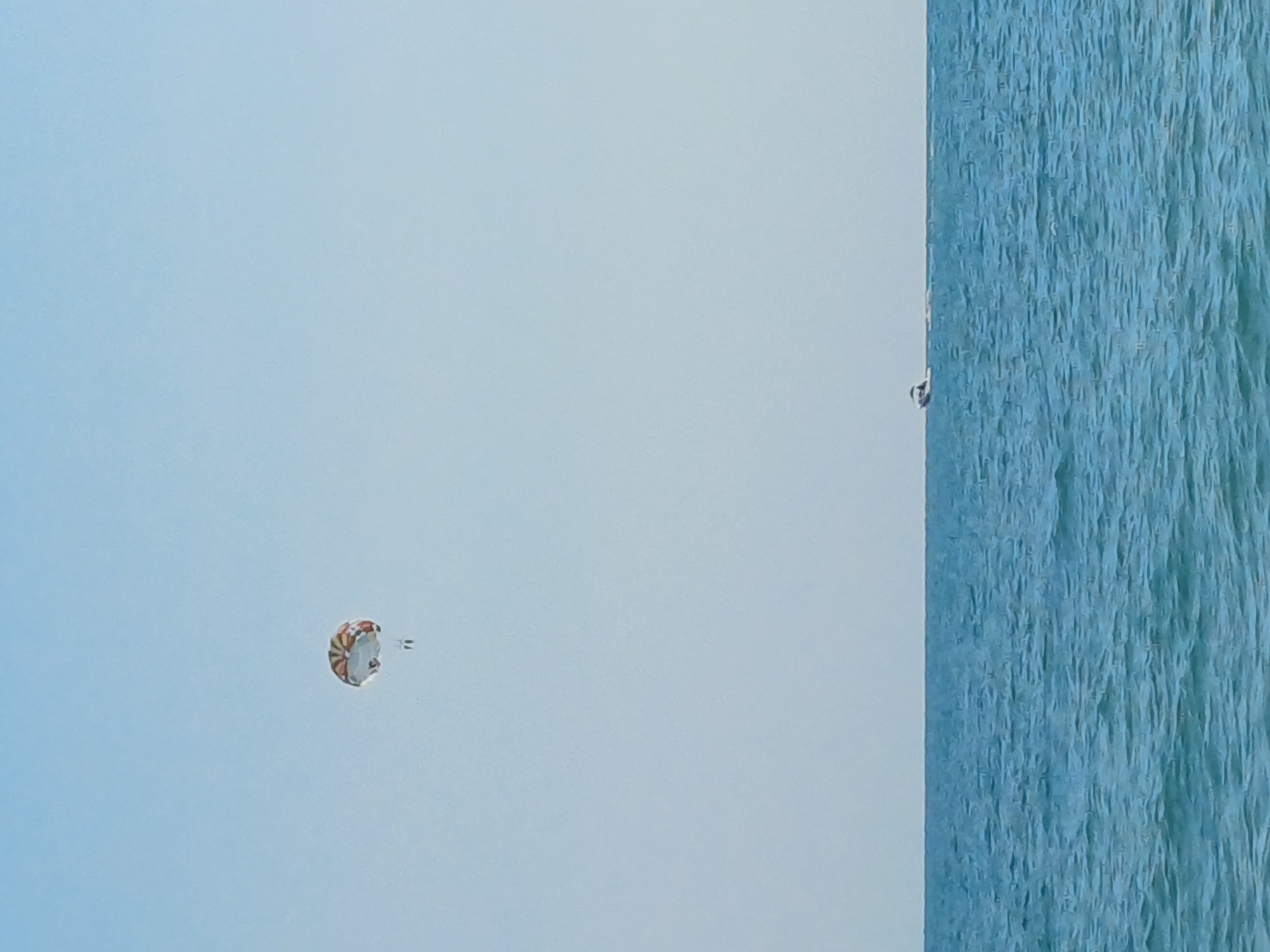 The image shows a clear blue sky above a calm blue sea. In the upper left portion of the sky, a colorful parachute is visible, with two people suspended beneath it. Towards the horizon, a small white boat is moving across the water, leaving a slight wake. The scene appears to be a sunny day at the beach, likely in Sochi, Russia, given the presence of parasailing and boats.