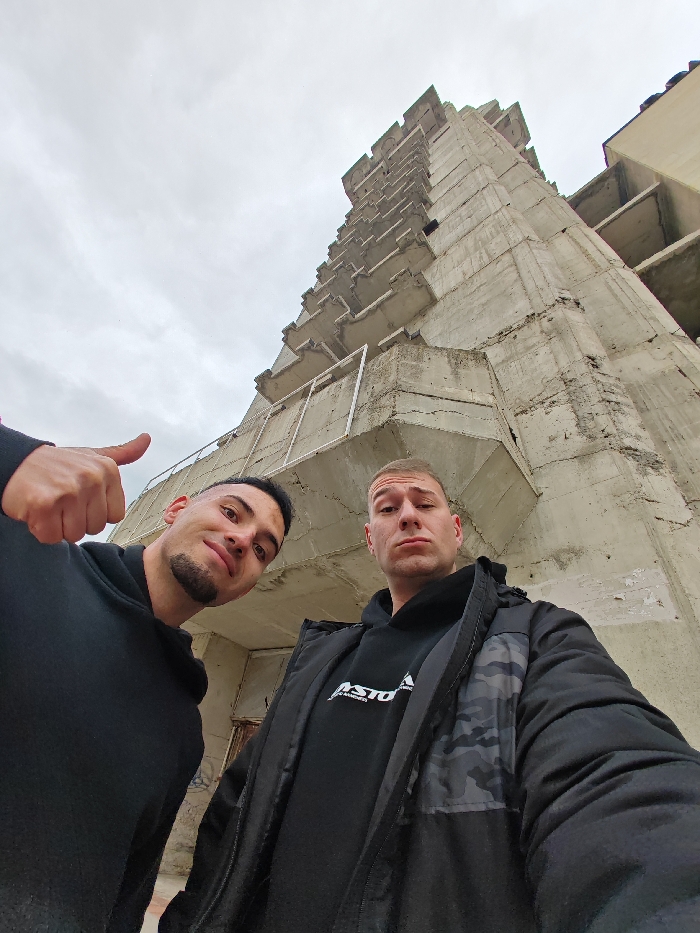 The image displays two adult males posing for a close-up photograph in front of the unfinished and abandoned Monument to 1300 Years of Bulgaria, situated on the Shumen Plateau, near Shumen, Bulgaria. The background sky is overcast.
The male subject on the left, with dark hair and a short beard, is wearing a black hooded sweatshirt. He exhibits a faint smile and extends his right hand with a "thumbs up" gesture. The male subject on the right, with light hair, wears a black hooded sweatshirt featuring a white "ASTON" logo, layered under a dark jacket with a grey camouflage pattern. His expression is neutral. Both individuals are looking directly at the camera.
The dominant background structure is a large, multi-story, brutalist concrete building, characterized by numerous cantilevered and stacked sections. The raw concrete surfaces show signs of weathering, and elements of its unfinished construction, including structural components, are visible. A white rectangular frame is present on a lower level of the building.