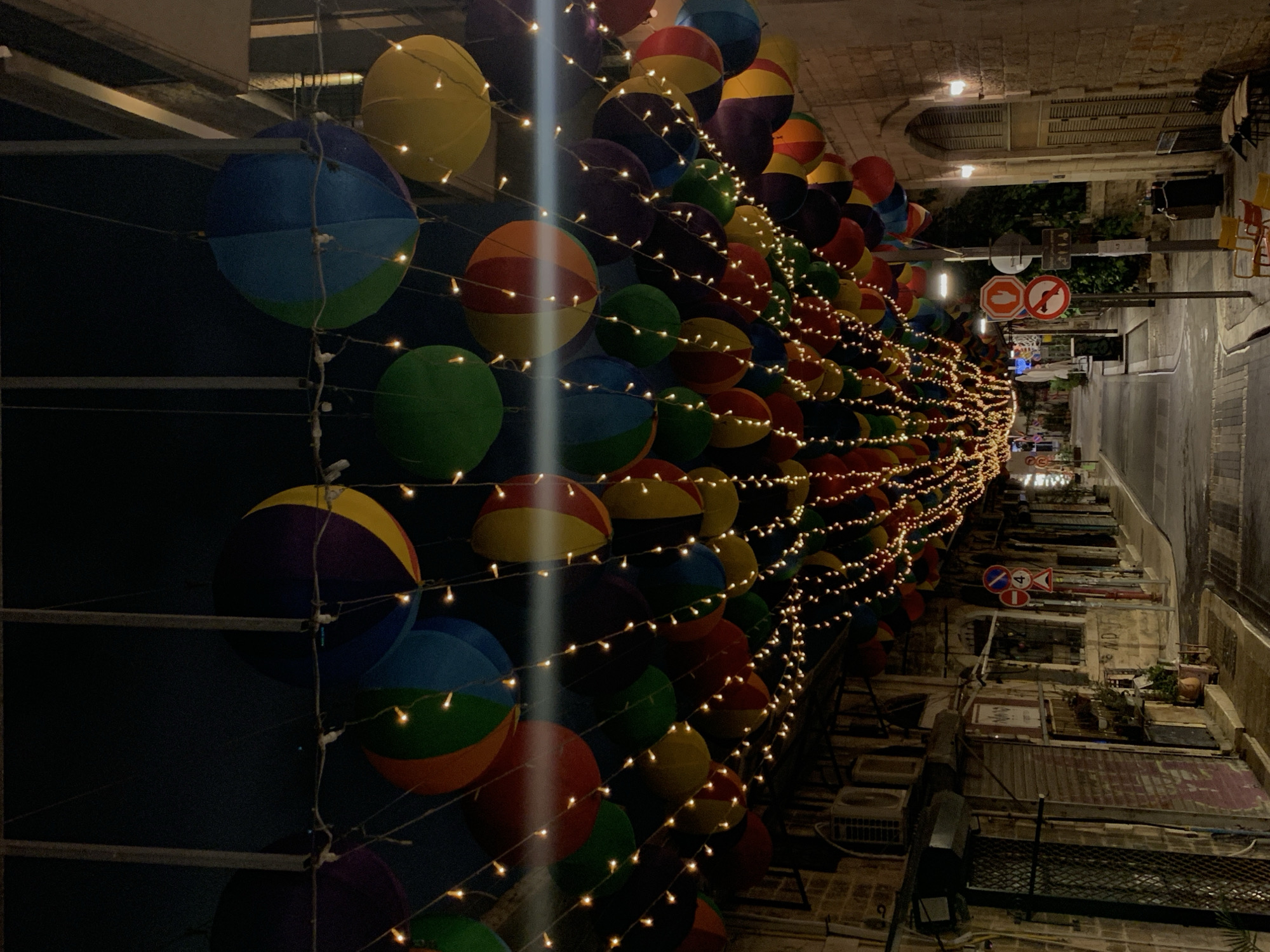 The image depicts a narrow street in Tel-Aviv, Israel, illuminated by strings of fairy lights and a canopy of colorful, round decorative objects, resembling opened umbrellas or balloons. These decorative items are suspended overhead, creating a vibrant and festive atmosphere. The street itself appears to be paved and wet, suggesting recent rain or cleaning.
On the left side of the street, buildings with traditional stone facades line the pathway. The structures feature closed shutters on windows and doors, and several air conditioning units are visible. Graffiti can be seen on some of the walls, and potted plants add touches of greenery.
On the right side of the street, more stone buildings are present, with arched doorways and shuttered windows. Several street signs are visible, including a stop sign and a sign with a number "4" inside a circular frame. Chairs and tables are set up outside, indicating a potential seating area for a cafe or restaurant, though no patrons are visible.
The overall scene is captured at night, with the artificial lighting from the string lights and building entrances providing the primary illumination. The dark sky above suggests it is late evening or night. The image is taken from a low-angle perspective, looking down the length of the street.