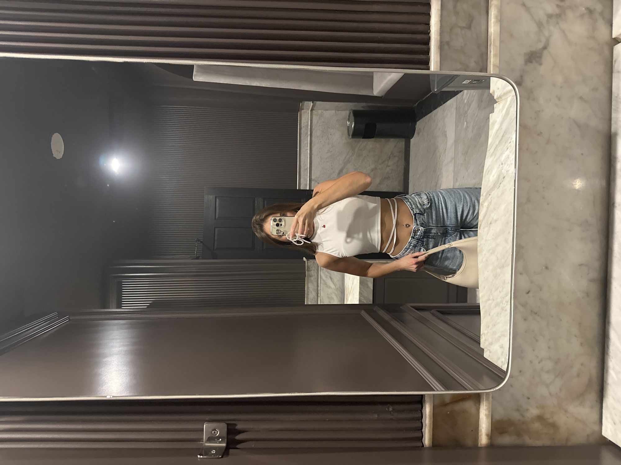 A woman stands in a dimly lit, modern public restroom, taking a mirror selfie. She is wearing a white cropped tank top with white string accents, light-wash denim shorts, and a silver belly button ring. She holds a light-colored purse in her left hand. The restroom features dark brown fluted walls, marble accents, and a black trash can. The lighting is low, with a bright overhead light reflected in the mirror. The overall aesthetic is sleek and contemporary, potentially located in a trendy establishment in North Kuta, Indonesia.