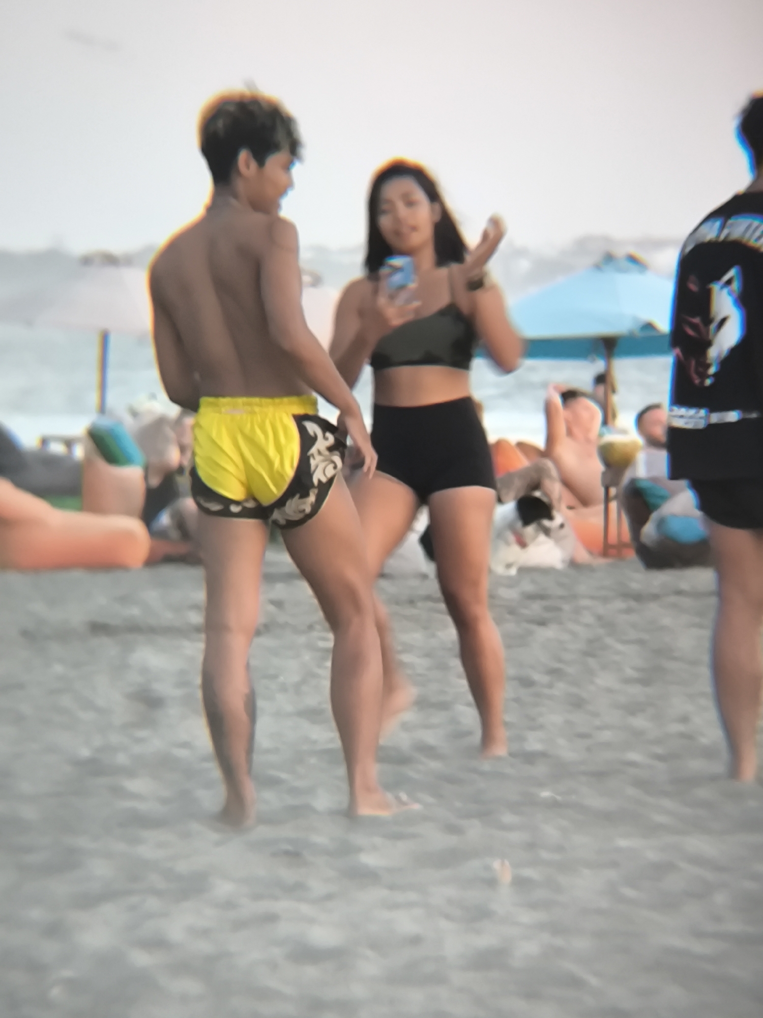 The image shows a beach scene in North Kuta, Indonesia. In the foreground, a shirtless individual with yellow and black Muay Thai shorts stands with their back to the camera. To their right, a person in a dark green sports bra and black shorts holds a phone. Further to the right is another person wearing a black t-shirt with a white graphic. In the background, there are people lounging on beach chairs and under umbrellas. The sand is gray.