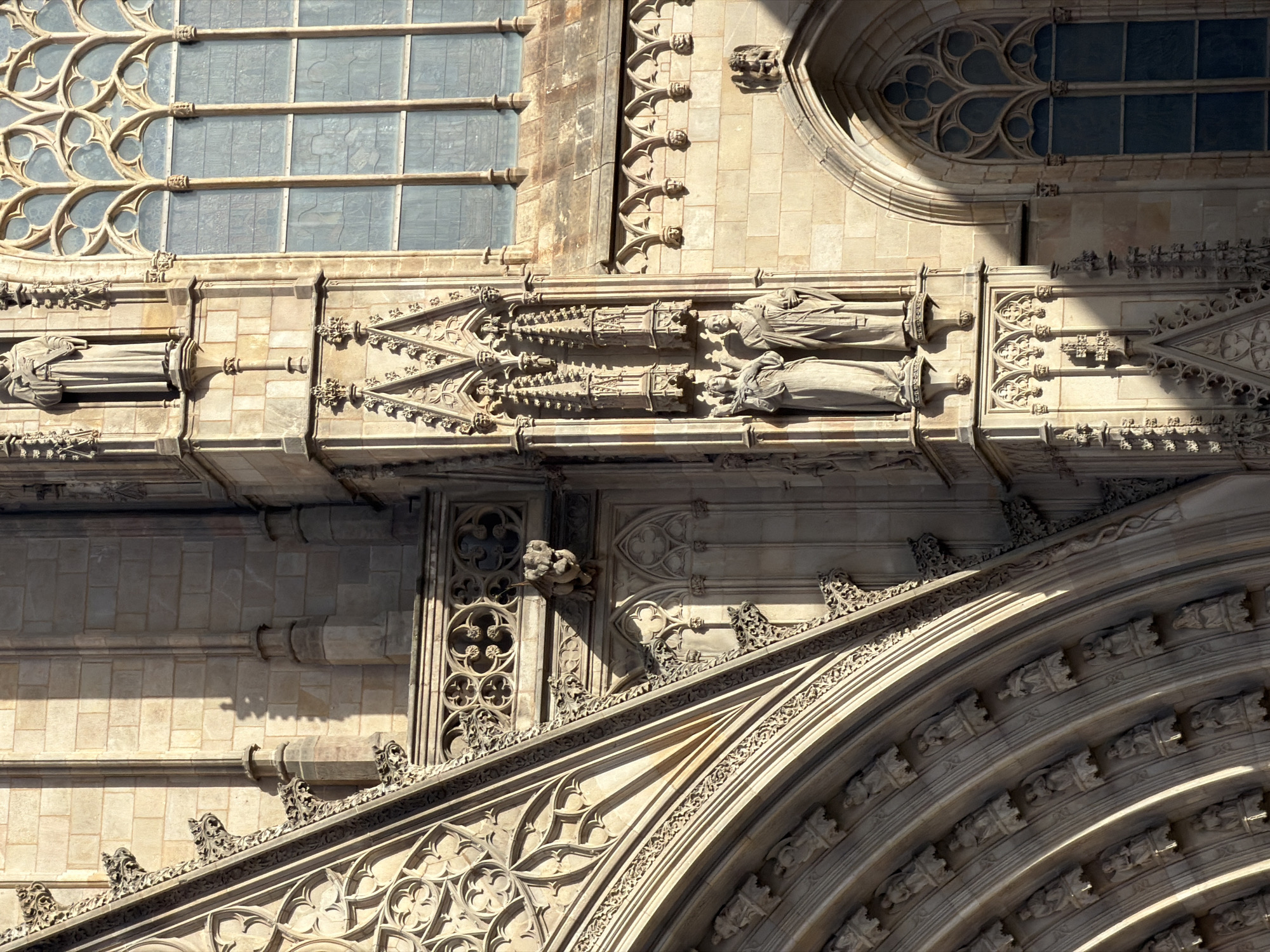 This is a close-up, eye-level, outdoor, daylight shot of the exterior of a Gothic cathedral in Barcelona, Spain, likely the Barcelona Cathedral (Catedral de Barcelona) given its architectural style and location. The focus is on intricate stone carvings and architectural details.
In the foreground on the left, a large, arched section of the cathedral is visible. This arch is composed of multiple concentric layers, each adorned with rows of small, carved figures or statues, appearing to be angels or saints, set within ornate niches. The stone is a light beige color, and the shadows highlight the depth and detail of the carvings.
To the right of the arch, the main facade of the cathedral rises. This section features more elaborate stonework, including pointed arches, spires, and decorative tracery. Two prominent statues are positioned in niches at mid-level. The statue on the left depicts a female figure with flowing robes and a headdress, holding a staff or rod. The statue on the right is of a male figure, also in robes, with a similar headdress and holding a cross or staff. Above these figures, another niche contains a standing statue of a robed figure, possibly a monk or saint, with hands clasped.
Further to the right, a large Gothic window with intricate tracery is visible. The window is divided into multiple panes, filled with a muted blue-tinted glass. Below this window, a series of smaller, arched niches are carved into the wall, each containing a small, carved face.
A prominent gargoyle or grotesque figure is visible on a ledge between the arched entryway and the main facade, casting a shadow on the wall. The overall impression is of a highly detailed, ancient, and grand religious structure bathed in the warm light of the sun, creating strong contrasts between light and shadow.