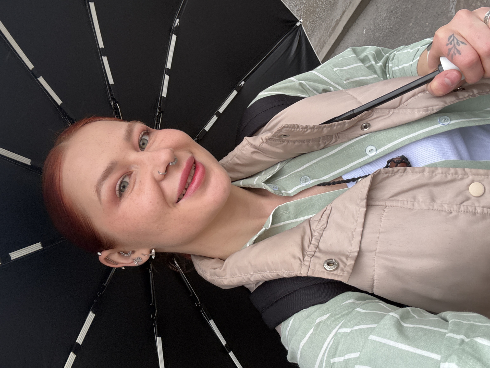 A young woman with freckles and multiple piercings in her left ear is holding a black umbrella. She is wearing a light green and white striped shirt underneath a beige vest. Her hair is reddish-brown. She has a nose ring and a tattoo on her hand. The image is taken at a close angle, looking up at the woman. The city location of Rostov-on-Don, Russia, cannot be confirmed from this image.