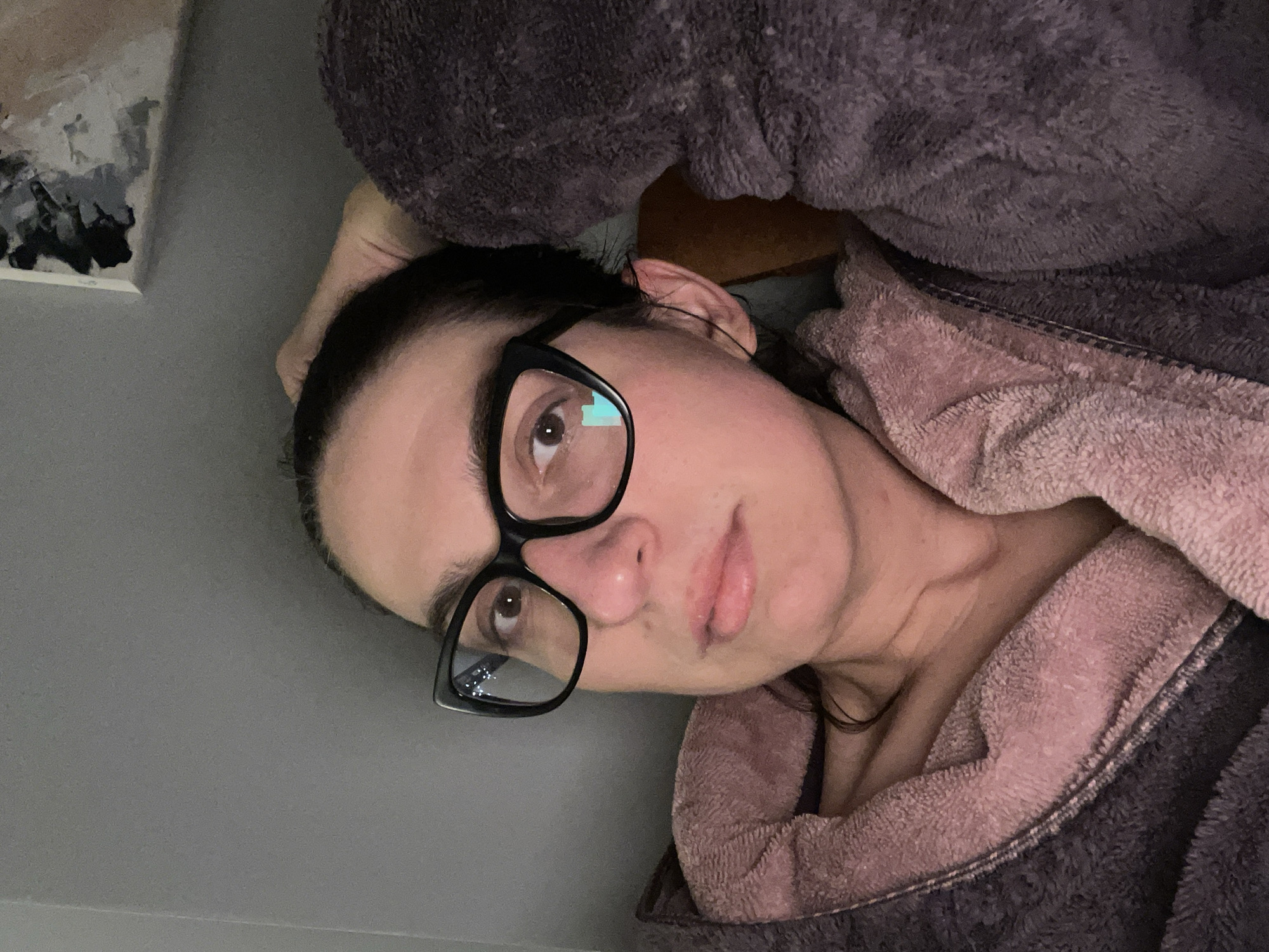 A woman with dark hair pulled back, wearing black-framed glasses, is reclining in what appears to be a plush, two-tone bathrobe or blanket (possibly gray and pink). Her head rests on one hand. The background is a neutral gray wall. To the top left, a framed abstract painting is visible. The location in Sofia, Bulgaria is unknown.