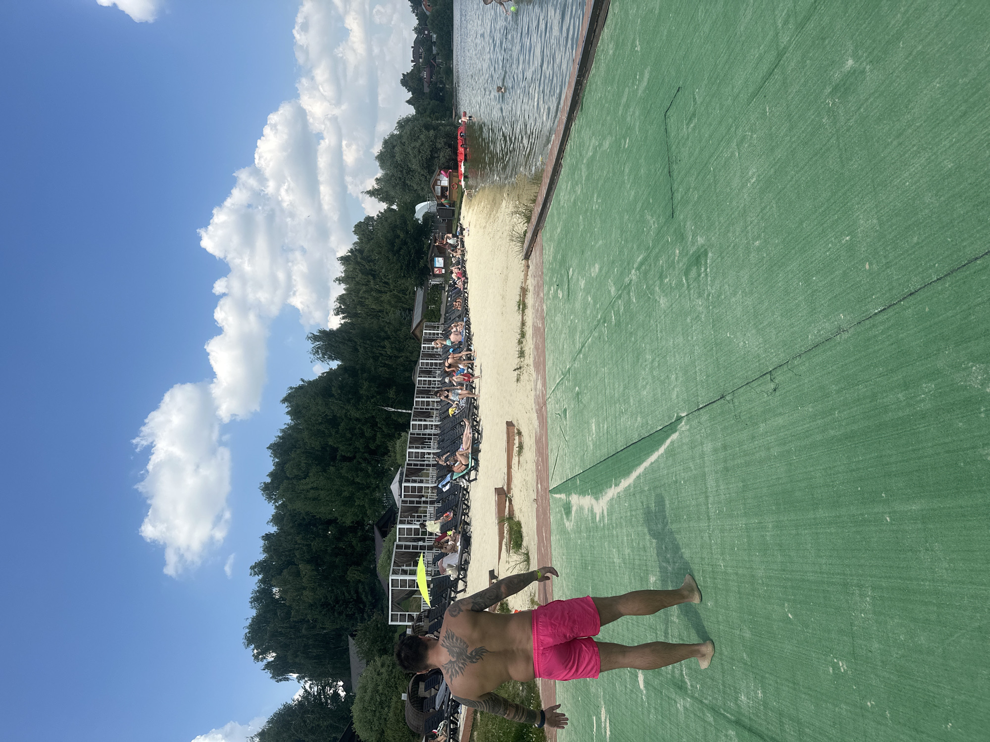 The image depicts a sunny day at a lakeside resort in Dal'nie Prudishchi, Russia. In the foreground, a man with extensive back tattoos and pink shorts stands on a green, textured surface, facing away from the camera. The surface appears to be a walkway or ramp leading towards the water. Behind him, a sandy beach is lined with rows of lounge chairs, many occupied by people sunbathing or relaxing. Further back, there are structures resembling cabanas and buildings, with a dense line of trees forming the backdrop. The lake is visible to the right, with several people in the water and some red pedal boats docked near the shore. The sky is blue with scattered white clouds.