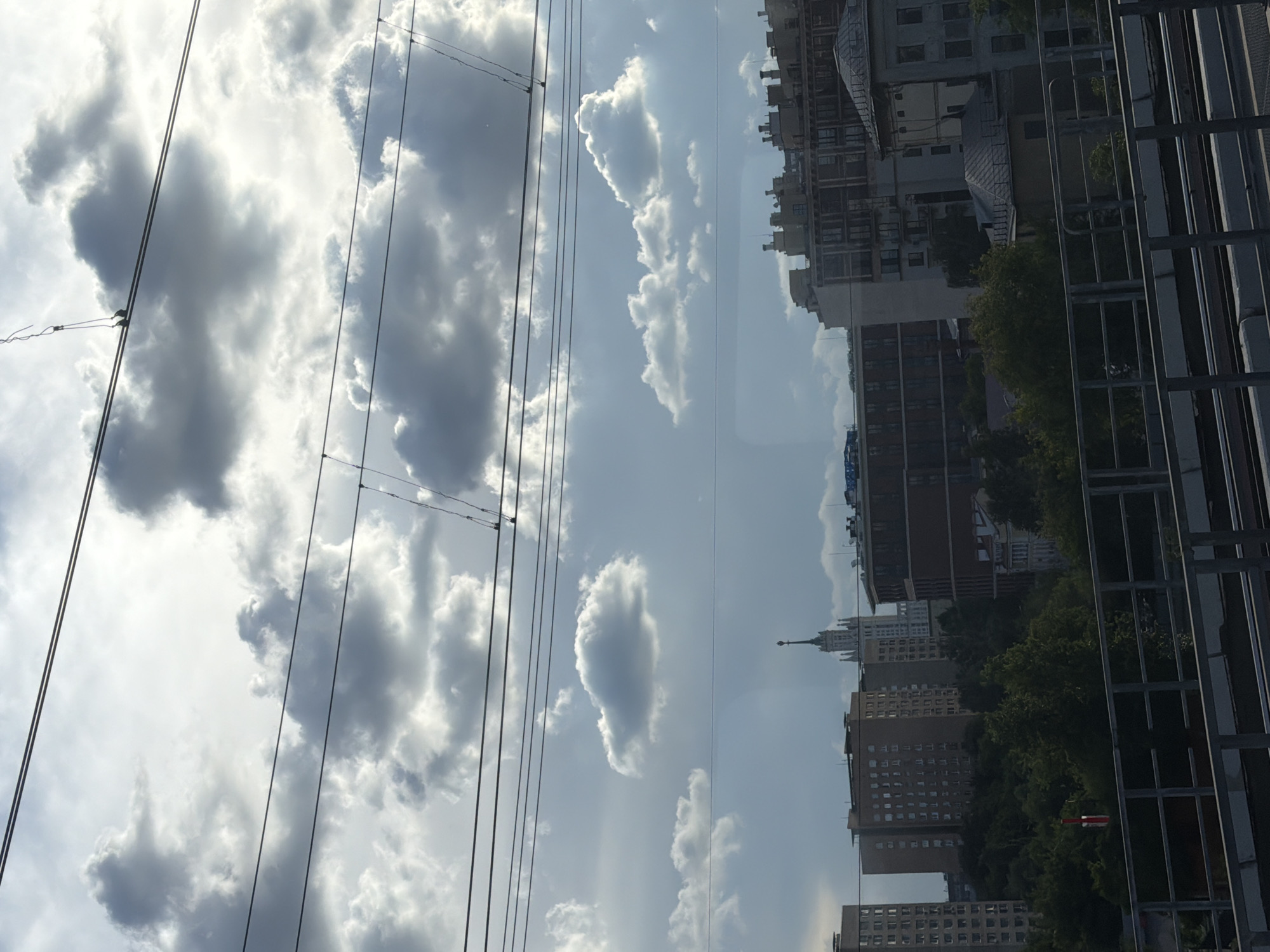 This is a photograph taken from a moving vehicle, likely a train, looking out at a city skyline under a cloudy sky. Overhead power lines are prominent in the foreground and mid-ground, crisscrossing the frame. The sky is filled with a mix of dark and light clouds, with patches of sunlight breaking through.
In the distance, a cluster of buildings forms the city skyline. Among them, a distinctive tall, slender building with a pointed spire stands out. There are also several mid-rise apartment buildings and what appear to be older architectural structures with multiple chimneys or decorative elements on their roofs. Dense green trees partially obscure the lower portions of the buildings, suggesting a park or green space within the urban landscape.
A railing and tracks, likely from the vehicle the photo was taken from, are visible in the bottom portion of the image. The overall impression is a view of a city during daylight hours, possibly during a break in a storm or on an overcast day. The location is identified as Unknown, Unknown, indicating that specific geographical details cannot be determined from the image.