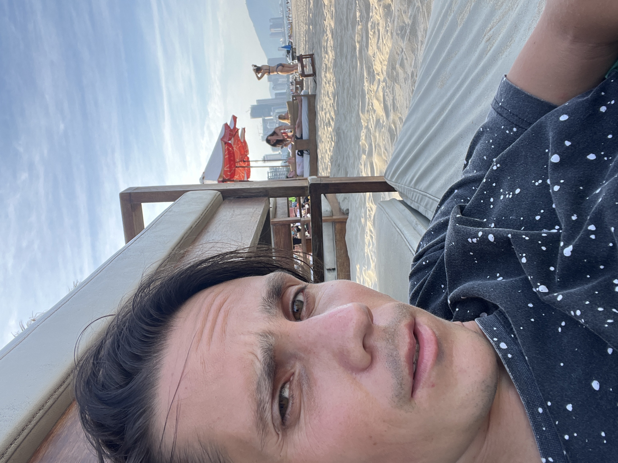 In Đà Nẵng, Vietnam, a man is captured in a close-up selfie on a beach. He is wearing a dark t-shirt with white speckles. In the background, the sandy beach stretches out, with several people relaxing on lounge chairs under umbrellas. Two women in swimwear are visible, one lying down on a lounge chair while the other stands. In the distance, buildings and mountains are seen against a bright, cloudy sky. The setting appears to be a beach resort area.