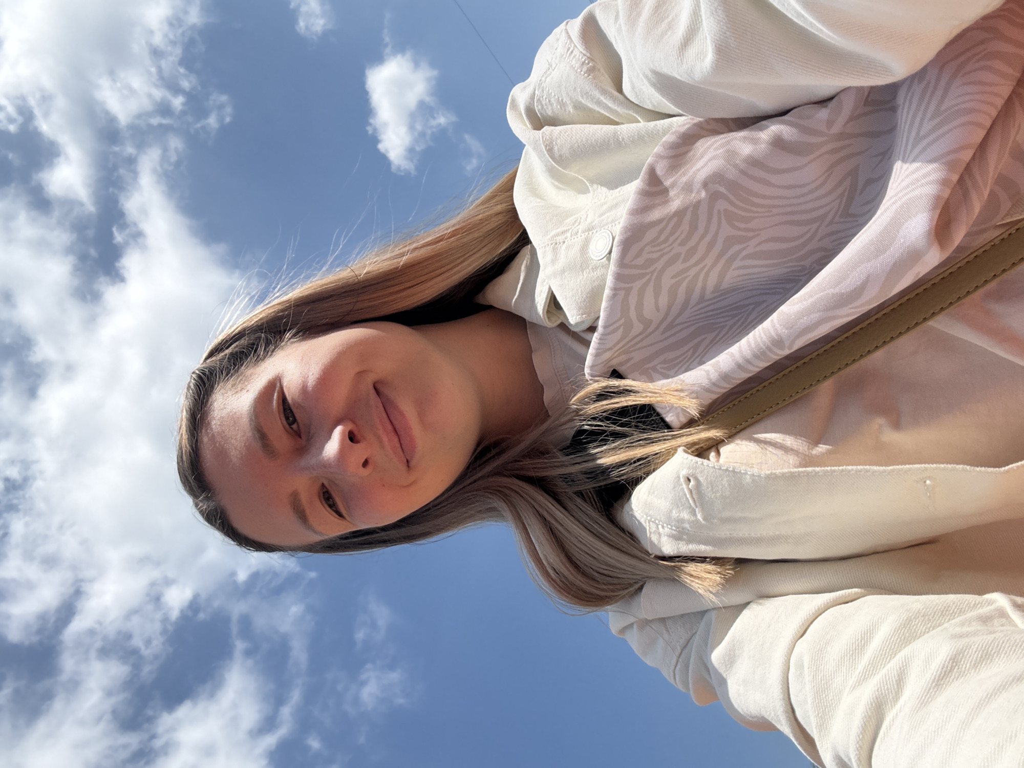 A low-angle, selfie-style shot captures a woman with fair skin and long, light brown and blonde hair smiling slightly at the camera. She is wearing a light-colored jacket and a patterned scarf. The background is a blue sky with scattered white clouds. There is no evidence to indicate this photograph was taken in Moscow, Russia.