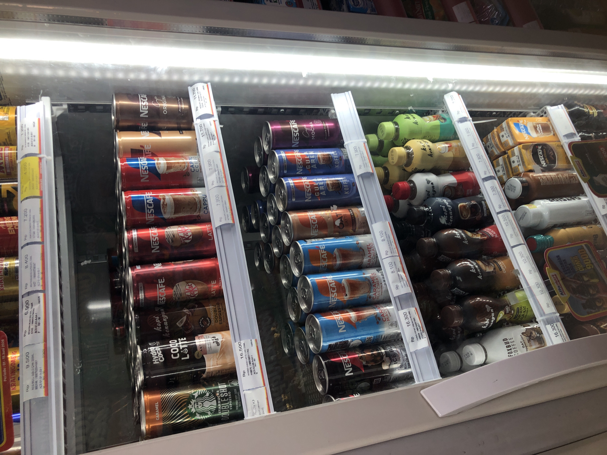 The image shows the inside of a commercial refrigerator, likely in a convenience store or minimarket in Putat, Indonesia, as indicated by the text "Alfamart" visible on a price tag. The refrigerator is stocked with various beverages, primarily canned coffee drinks and bottled coffee drinks.
On the top shelf, there are several rows of canned coffee. Brands visible include Nescafe and Hydro Coco. The cans come in various flavors such as Caramel Macchiato, Cappuccino, and Original. Some cans have red, blue, and brown labels.
The middle shelf also contains canned coffee, with many Nescafe cans. Some are labeled "Ice Blended," "Caramel Macchiato," "Gula Aren," and "Mocha." There are also some other canned drinks with gold and brown labels.
The bottom shelf holds bottled drinks. There are several white bottles that say "FOXBUD COFFEE OAT," and other brown and multi-colored bottles with text that appears to be Indonesian. Some of these bottles are branded "Kopi Kenangan" and "Kopi Susu Alpurat." There are also small yellow boxes of Nescafe Coffee Creamer.
Price tags are visible throughout the shelves, indicating prices in Indonesian Rupiah (Rp). The lighting in the refrigerator is bright and white, likely from LED strips, illuminating the products. The overall scene is neat and organized, suggesting a well-stocked retail environment.