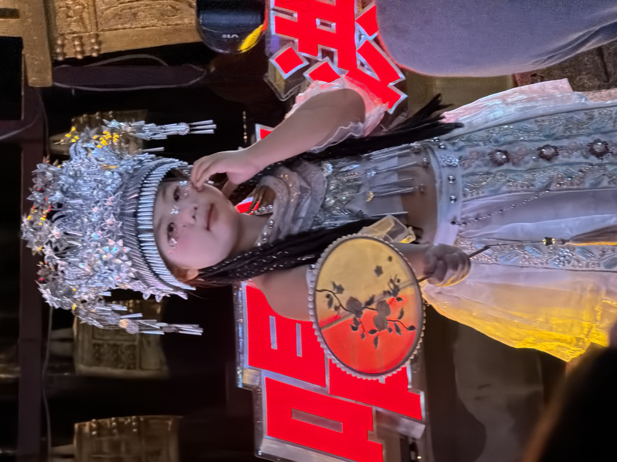A young girl, dressed in traditional Chinese attire and a sparkling headdress, stands on a stage. She holds a decorative fan in her hand and touches her face with her other hand. The background features large red Chinese characters and what appear to be ancient bells. The girl's costume includes a white skirt adorned with intricate patterns and beading, and a silver vest. Her makeup includes subtle glitter on her eyelids and cheeks, and rosy blush. The scene suggests a cultural performance, possibly in Yuzhong District, China, given the presence of traditional elements and Chinese characters. The lighting is dramatic, highlighting the girl and her costume against the darker background.