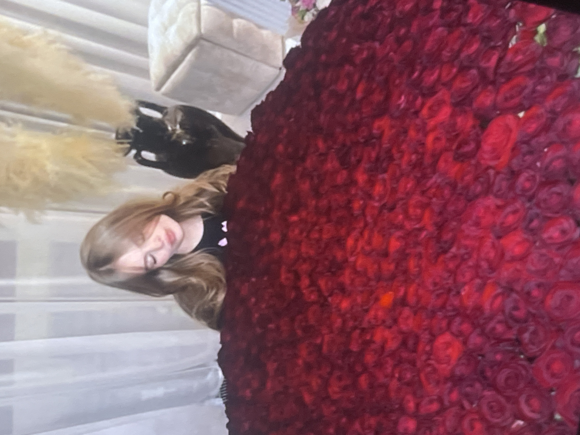 An indoor photograph captures a young girl alongside a voluminous arrangement of red roses. The girl, with long, light brown hair, is positioned on the left of the frame, her head slightly tilted. She wears a dark top with light-colored embellishments, possibly small hearts, visible near her shoulder. Her expression is soft and appears content, suggesting she is admiring the flowers.
The central and right portions of the image are dominated by a massive, dense display of deep red roses. The flowers are tightly packed, forming a rich, textured surface that fills a significant portion of the scene.
In the background, on the far left, soft, fluffy, light-colored decorative elements, possibly pampas grass, are visible. Above these, a light-toned, upholstered piece of furniture, such as a stool or ottoman, can be seen. The setting suggests a well-appointed indoor space, likely a home or venue in Poltava, Ukraine, implying a special occasion or a grand gesture. There is no visible text in the image.