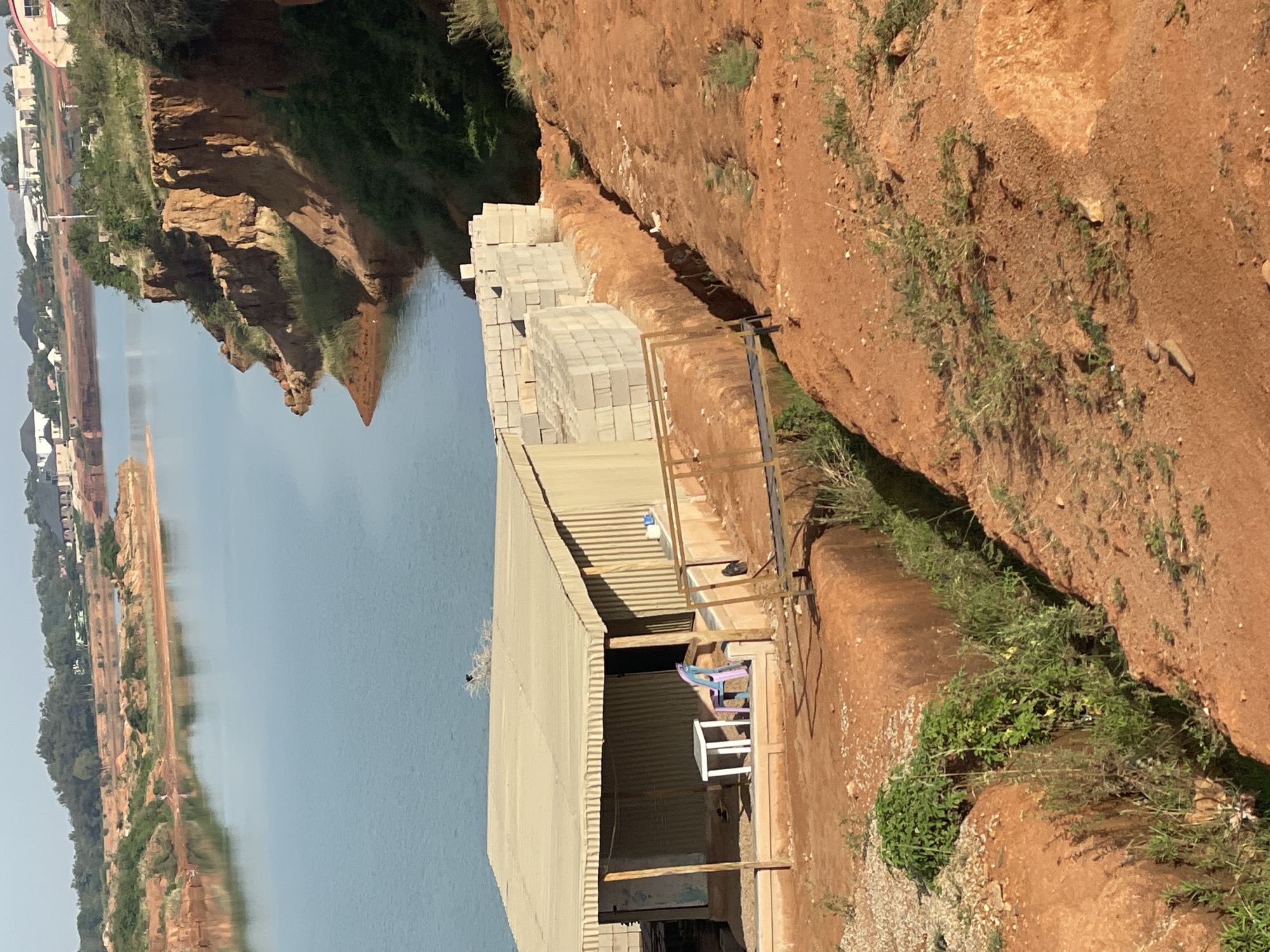The image shows a scene in Kwang, Nigeria, with a body of water to the left, possibly a lake or river. The opposite bank is visible, with structures and vegetation lining the water's edge. On the right, there is a steep, reddish-brown embankment. A metal structure resembling a shed is situated on the ground between the water and the embankment. Piles of gray concrete blocks are stacked near the edge of the embankment, close to the metal structure. Inside the shed, there are chairs and a table. The sky is visible as a sliver at the top of the image.